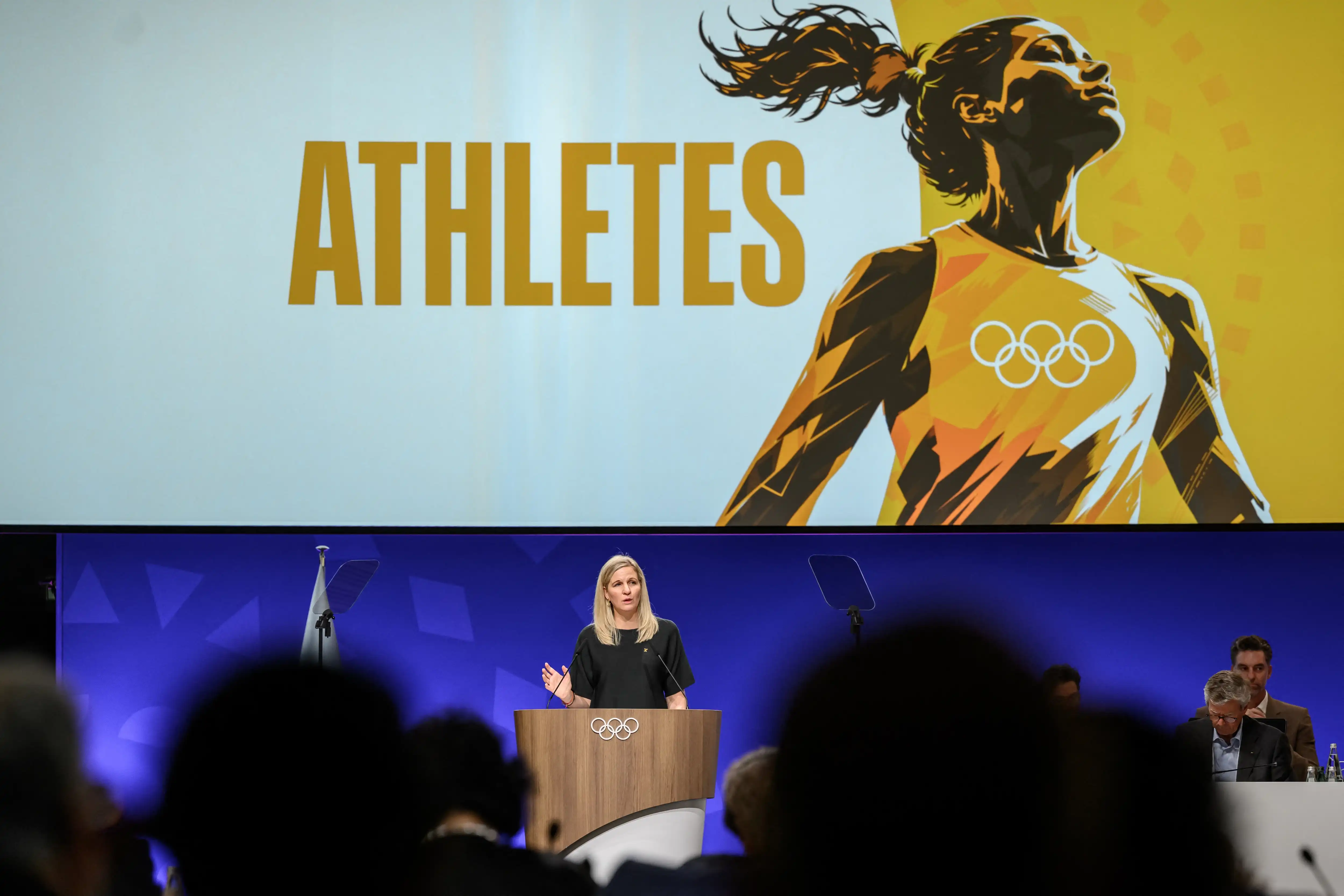The President of the International Olympic Committee delivers a speech in front of an animated banner of a female athlete