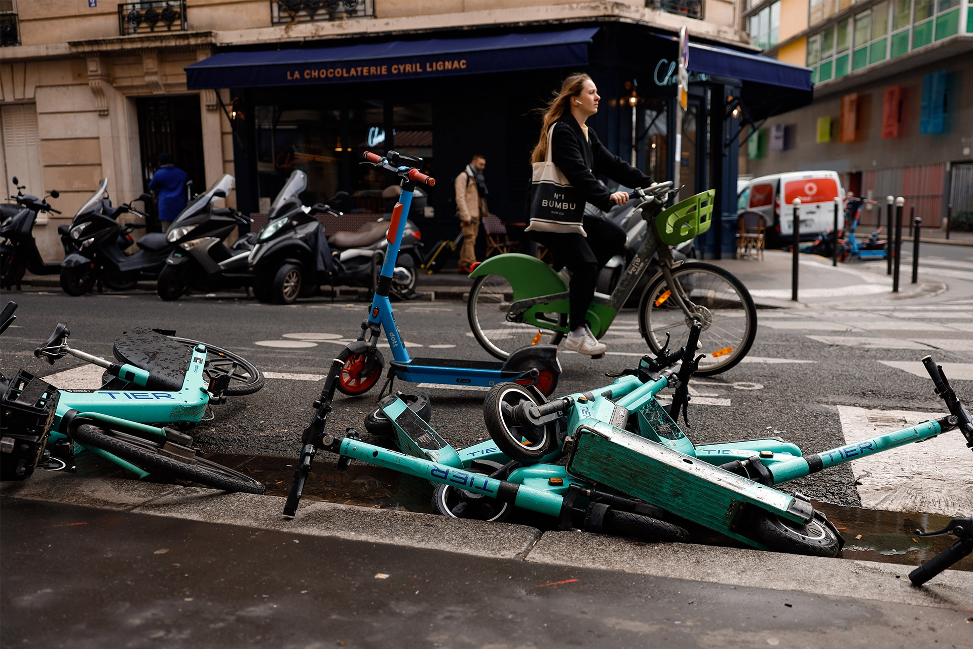 Woman riding an e-bike through Paris, with a pile of discarded e-scooters lying on side of road