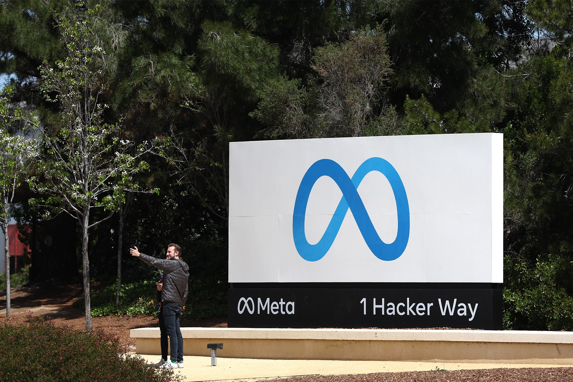 Person taking a selfie in front of large Meta sign, outside Meta headquarters in California