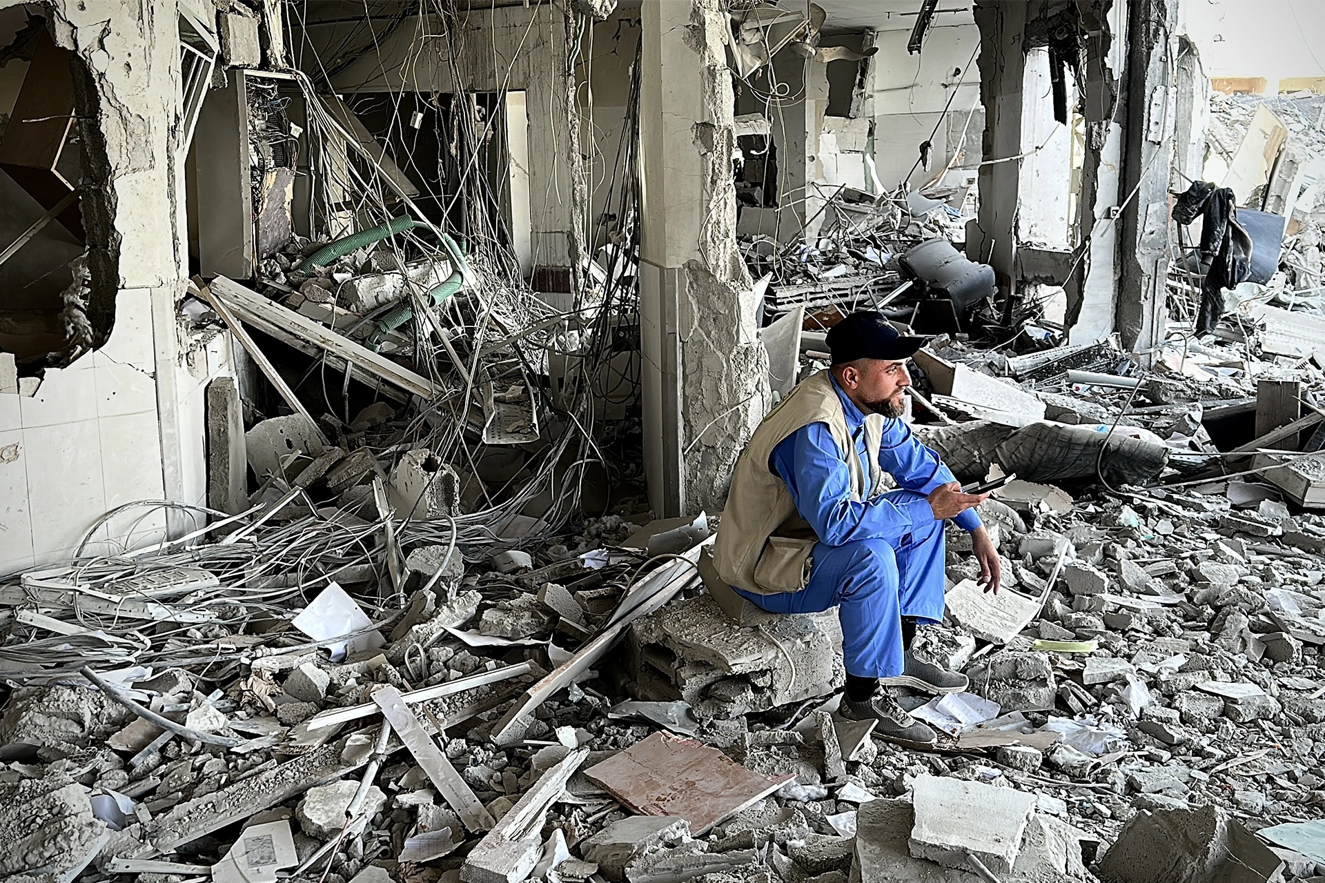 Man wearing blue hospital scrubs sitting in the shell of a bombed hospital building