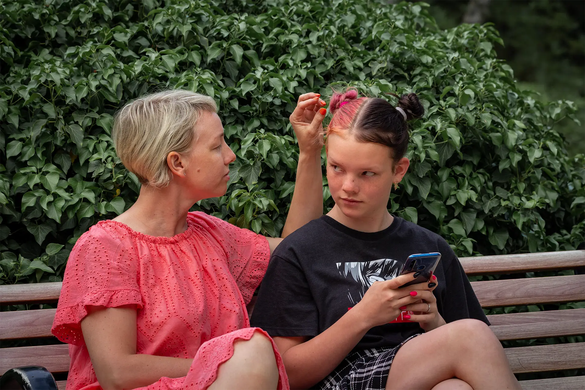 A child holding a phone looks at an adult, likely their mother, sitting next to them on a bench