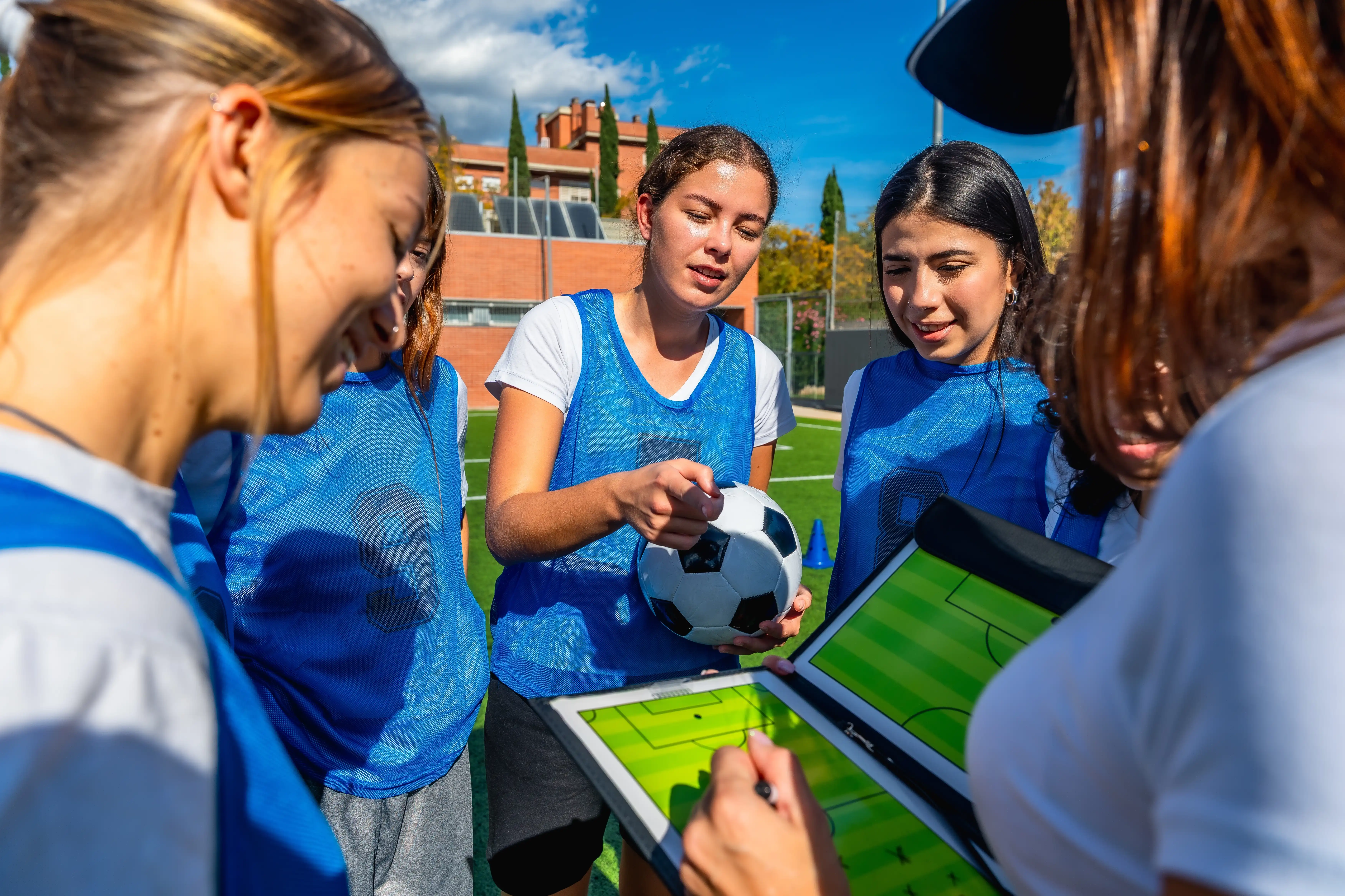 Young girls in blue bibs and sportswear point to a printed soccer pitch in a folder where a female coach is explaining a drill