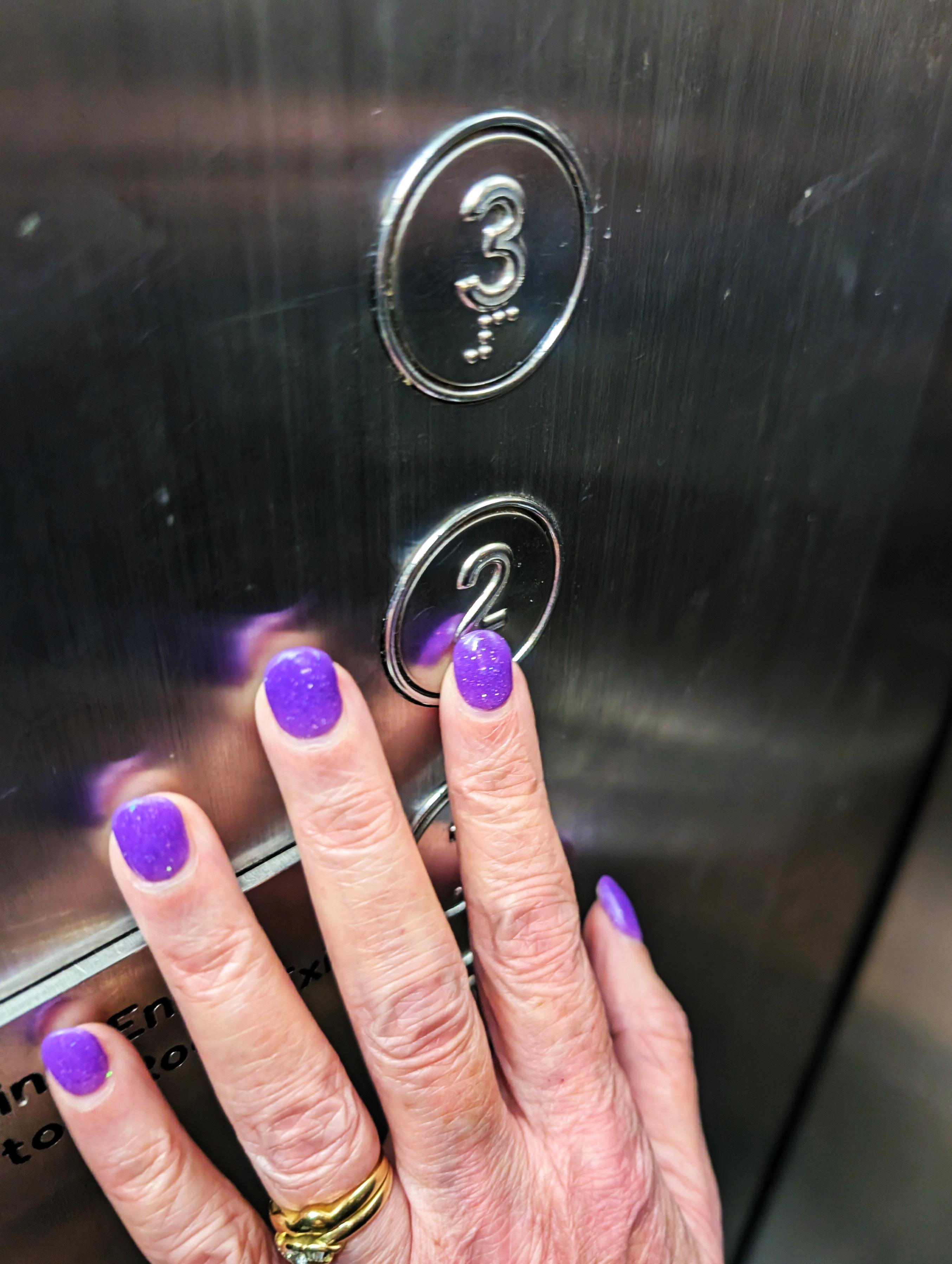 A hand with purple nail polish touching a the elevator button to read the braille.