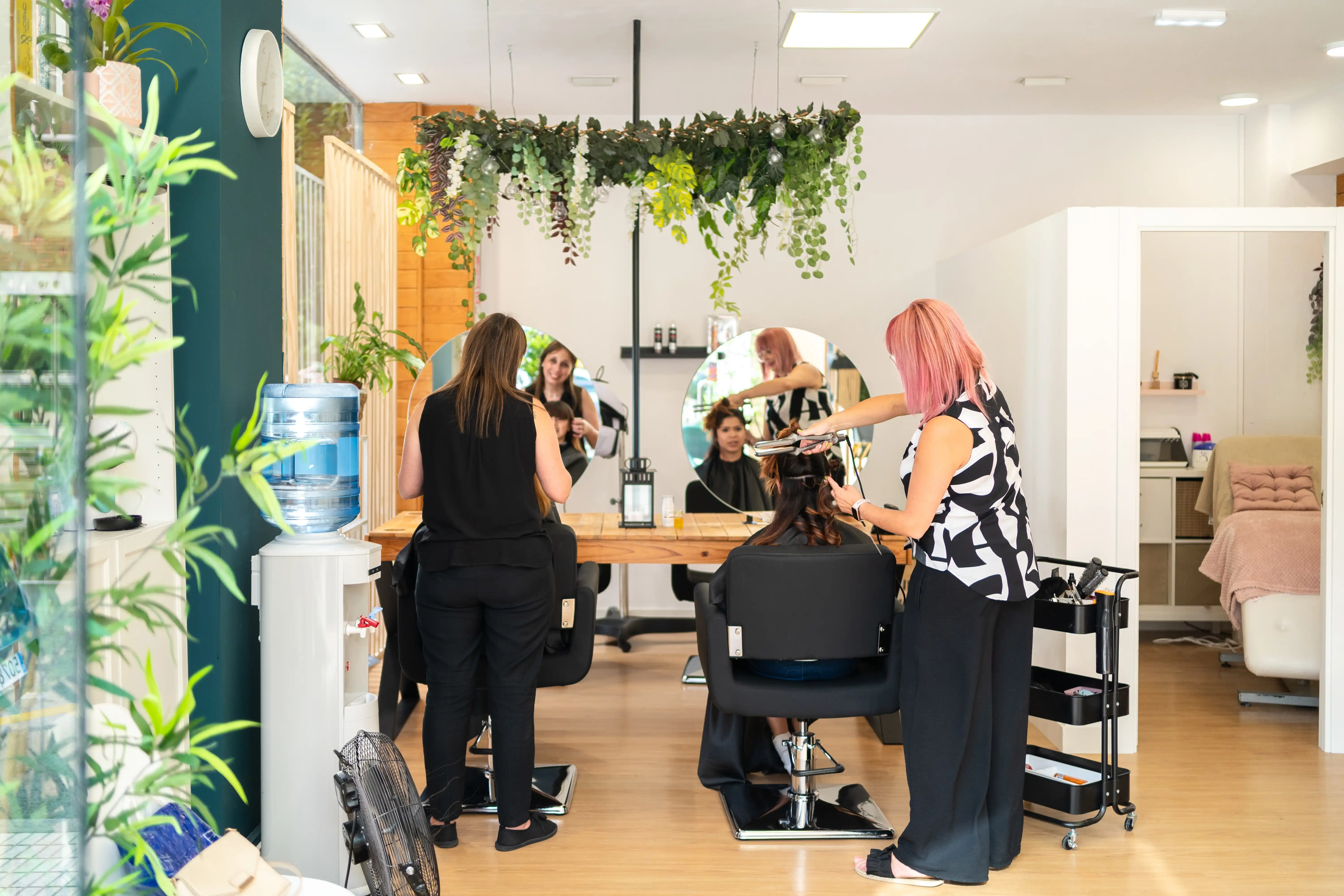 Two hairdressers stand side by side with clients in a chair who are facing a large mirror. The hairdressers are curling the clients hair with curling irons in a brightly lit beauty salon