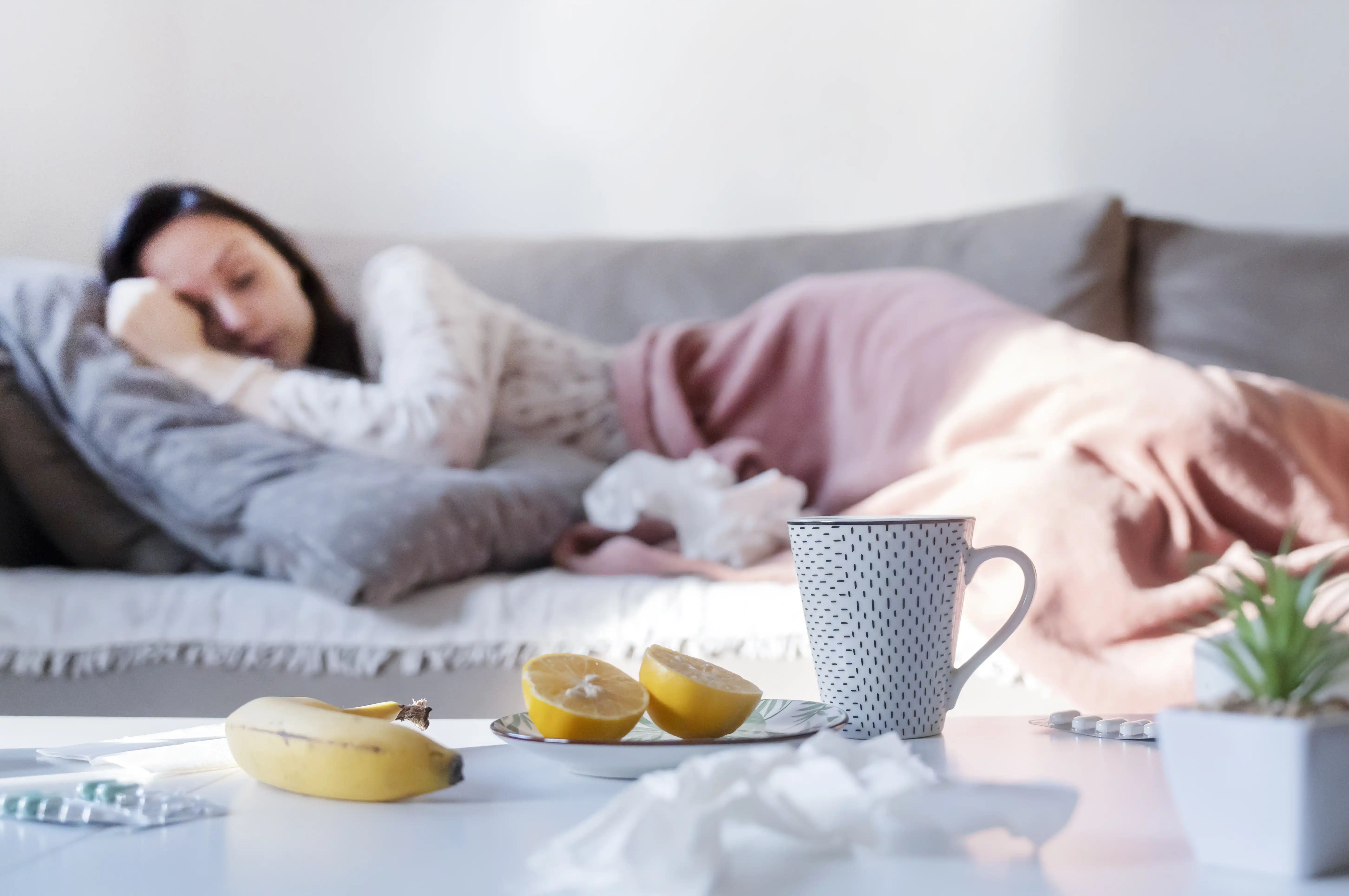 A woman rugged up on a couch with a mug of tea and banana on the coffee table in front of her