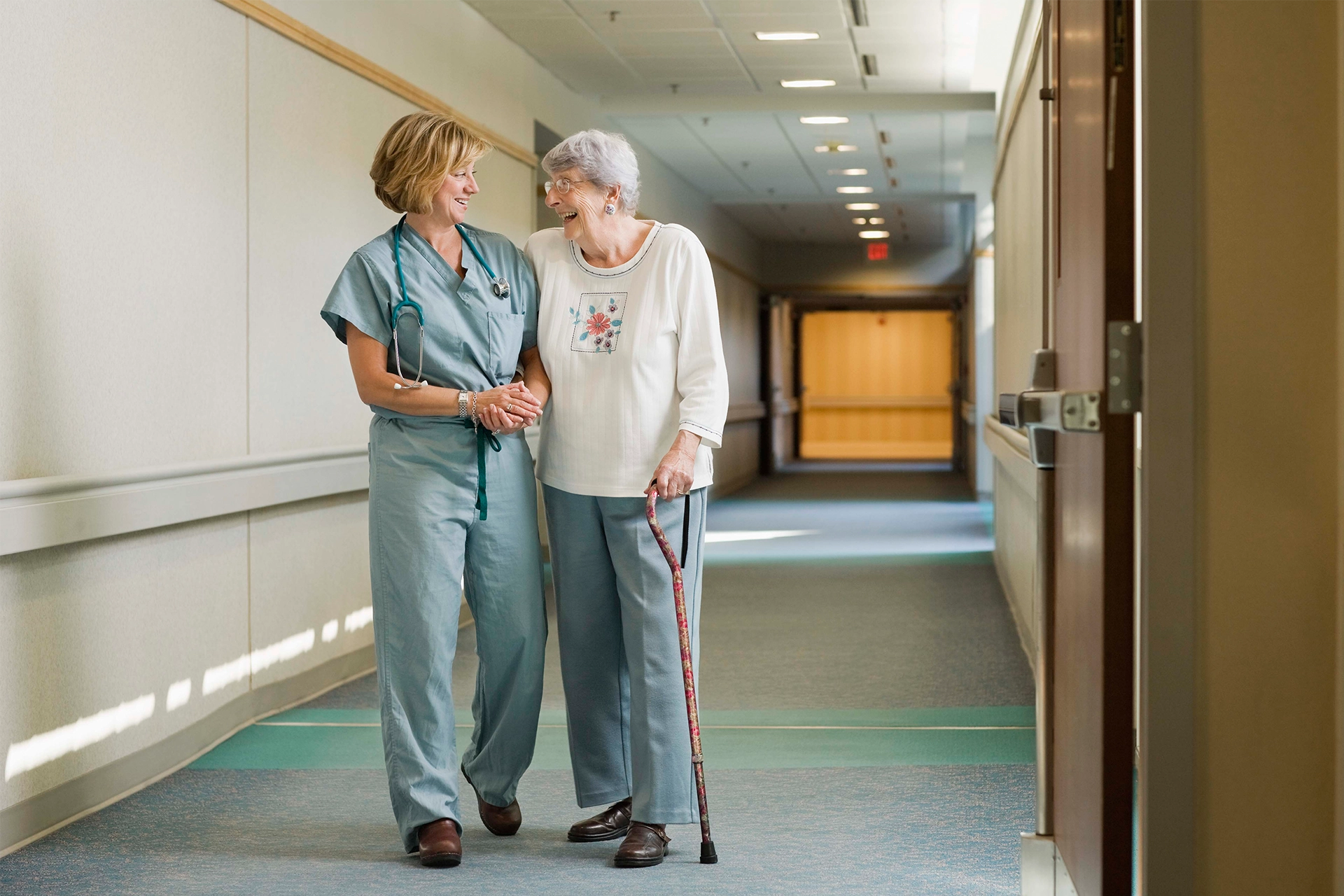 Middle aged nurse helping an helping woman walk down corridor in hospital, both are smiling