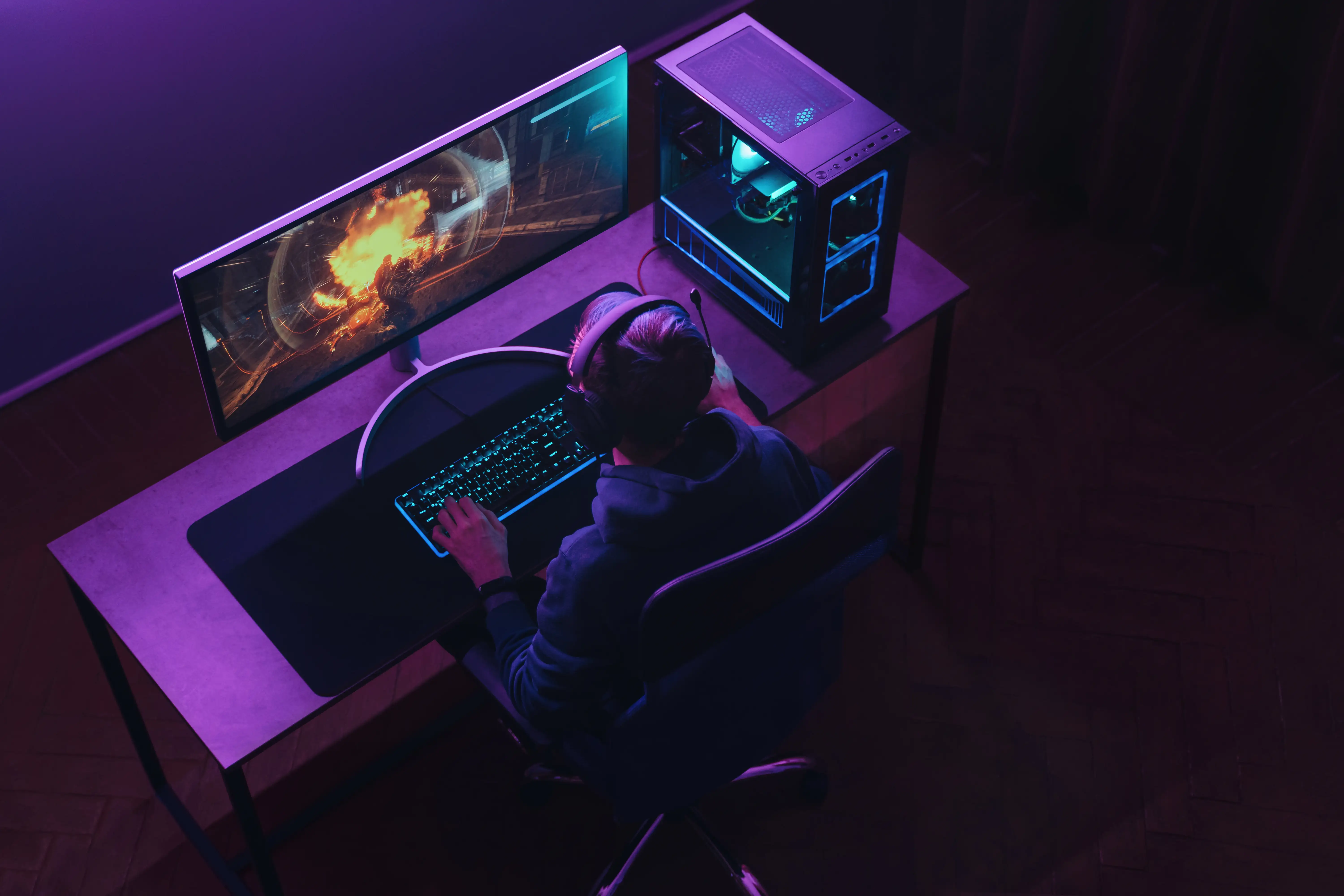 Overhead view of professional cyber sportsman in headphones playing shooter video game on his powerful gaming PC in dark neon room at night.