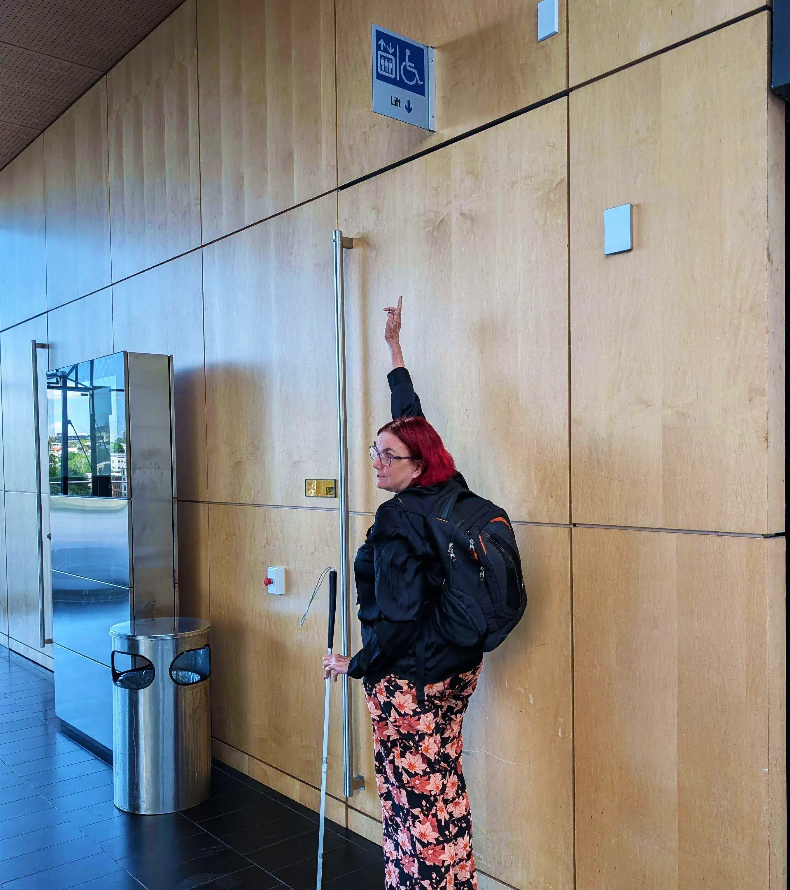 The panelled wall with a very hard to see sign indicating the lift is behind the door. Sheelagh, holding a cane, stands on her tip toes to point to the sign which is mounted very high above the door.