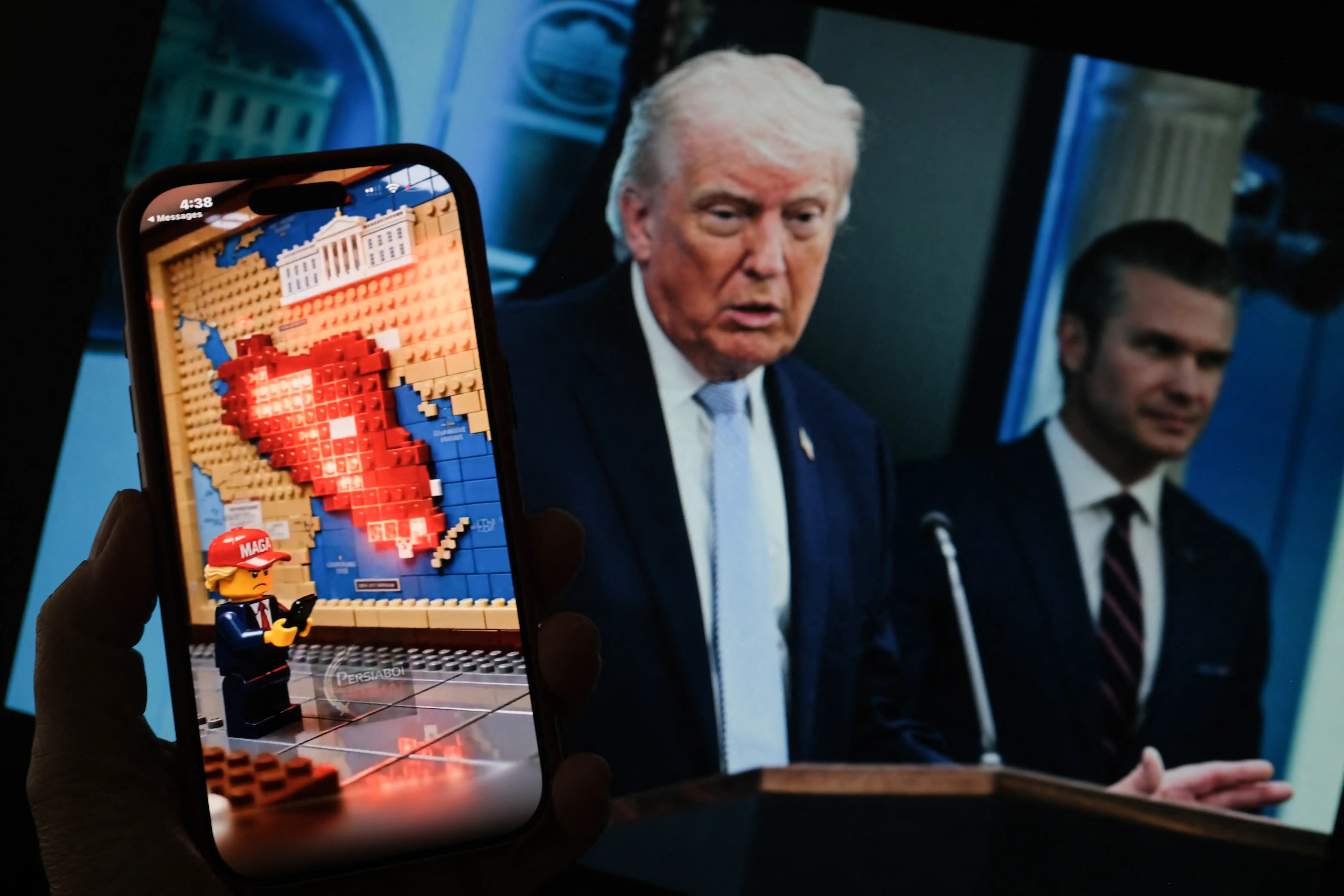 A Lego-style AI-generated war-themed video playing on a smartphone screen in front of a photo of US President Donald Trump and US Secretary of Defense Pete Hegseth. 