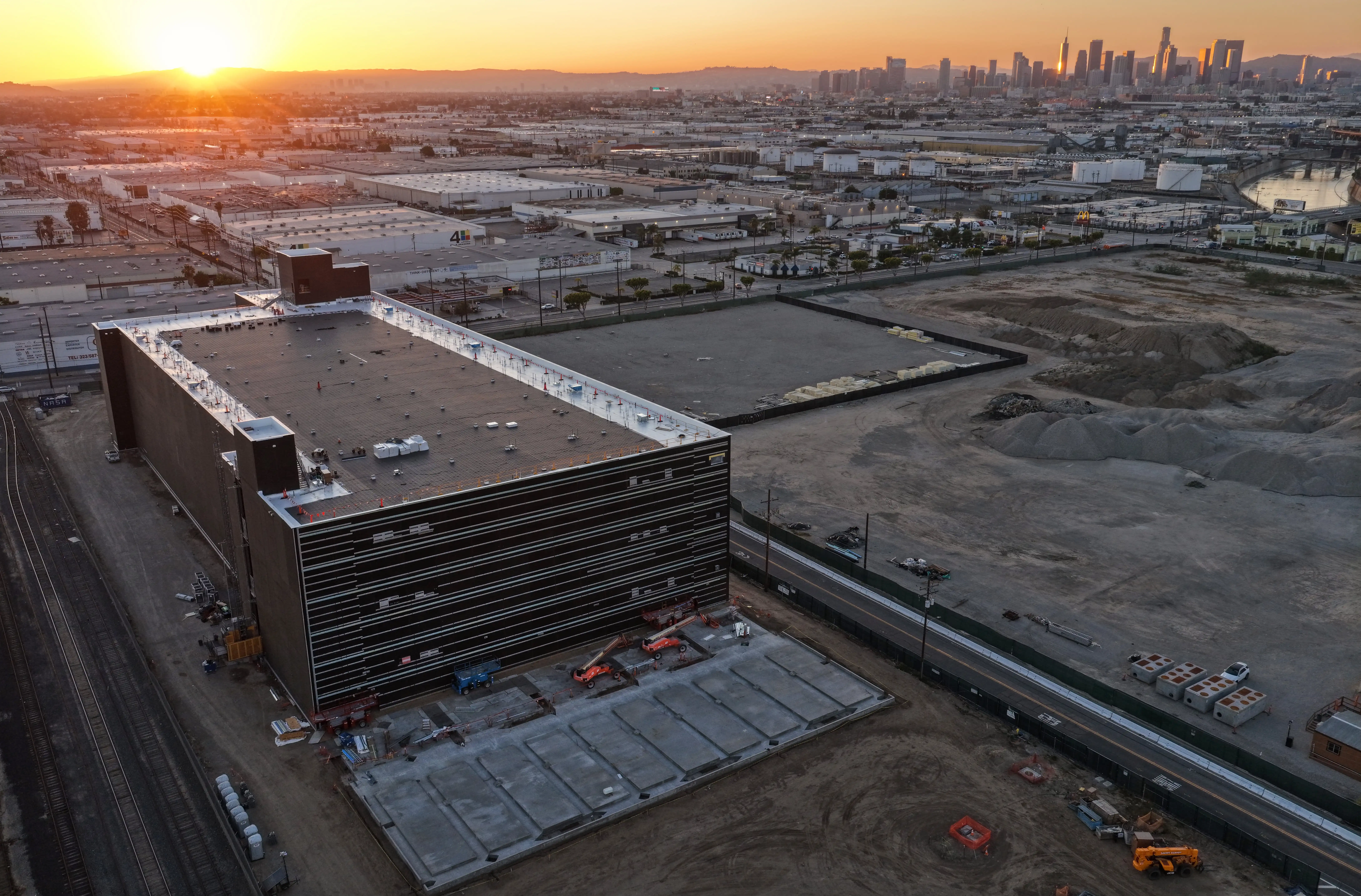 An aerial view of a new data centre being built. It is made of concrete and surrounded by large open spaces of dirt for future centres. The city is seen in the background and the sun is setting.