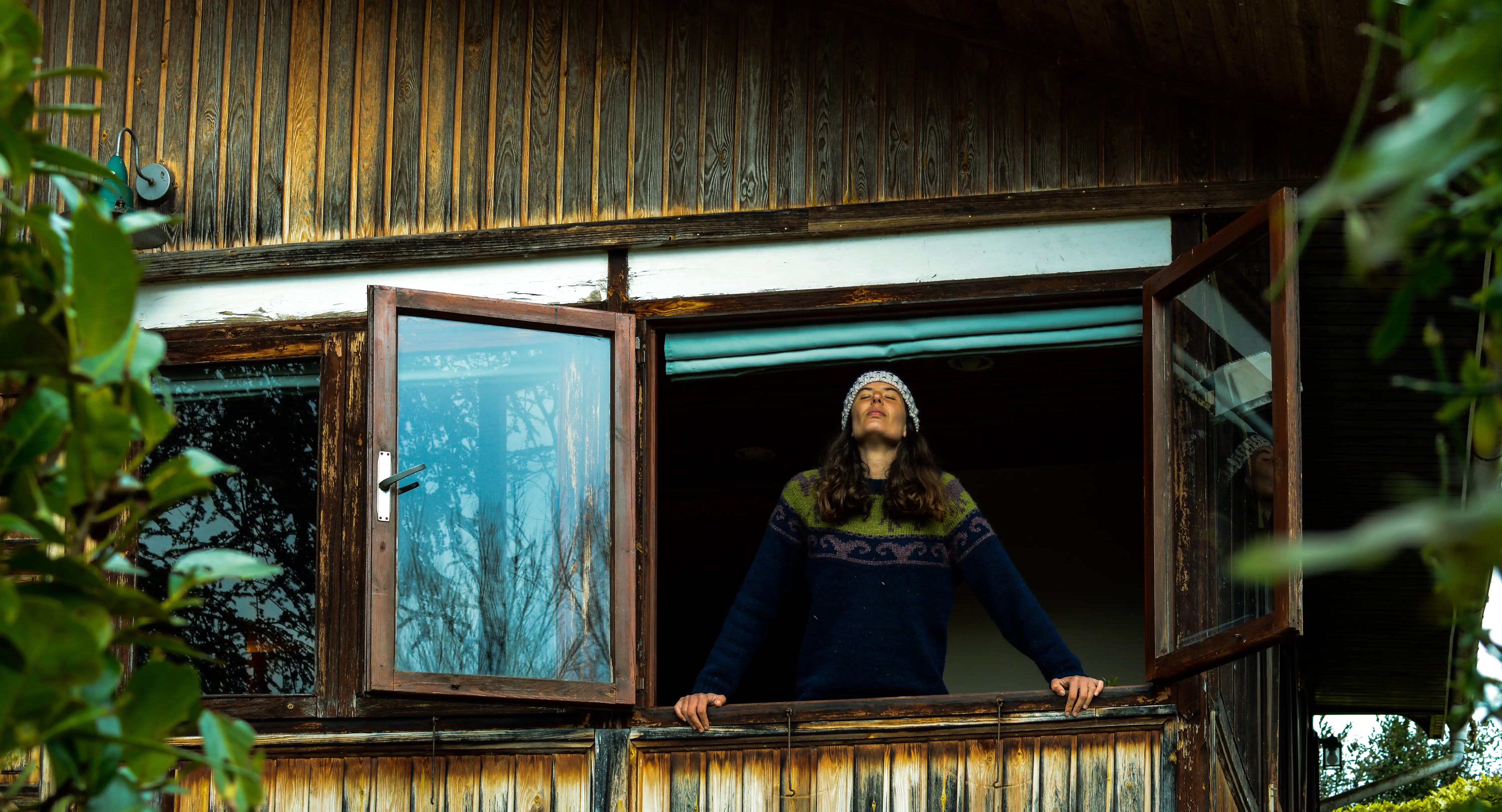 Women in a fleece jumper and beanie breathes in deeply through her nose looking out an open window in a winter cabin