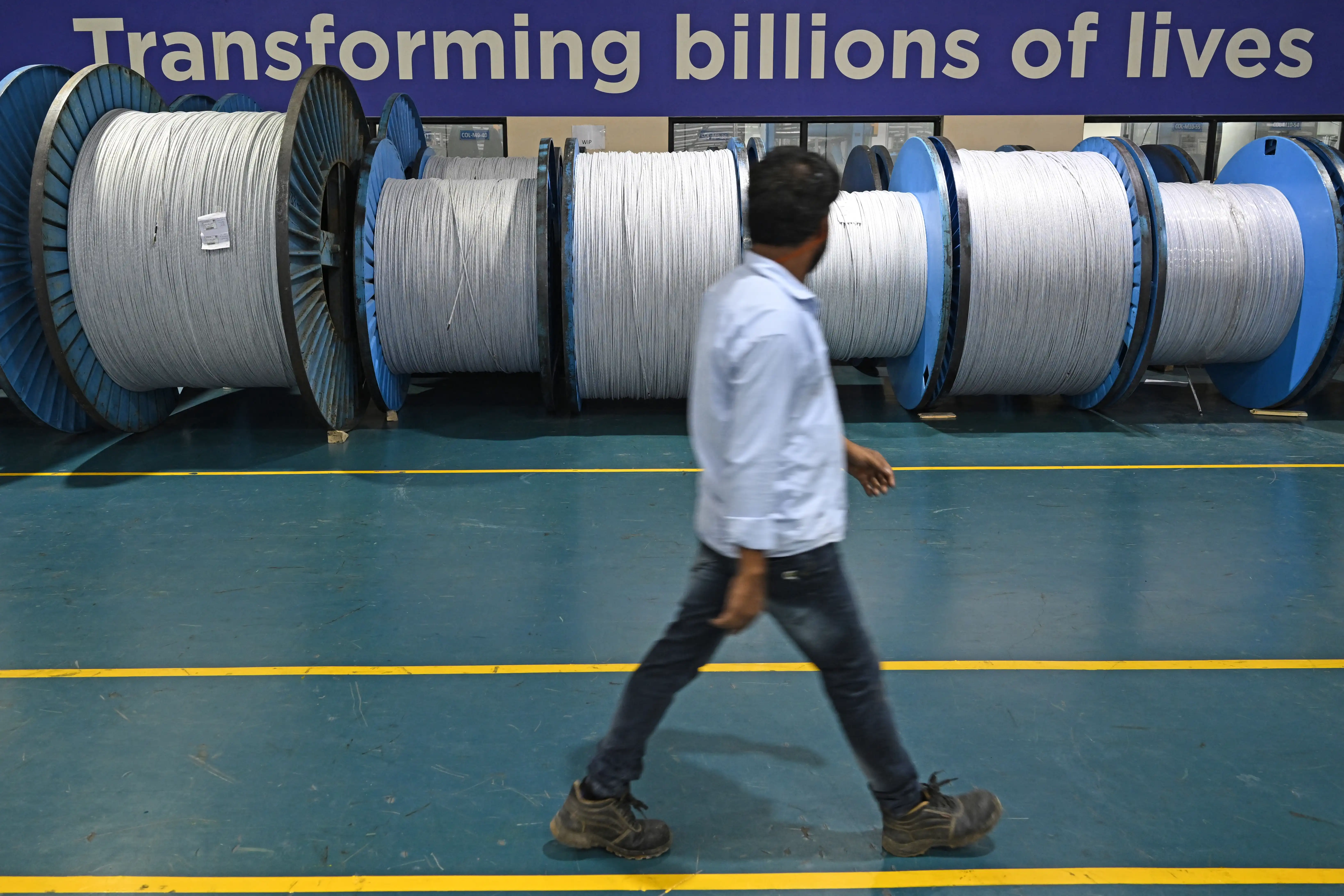 A man walks in front of huge spools of optical fibre cables with a sign that reads ‘Transforming billions of lives’