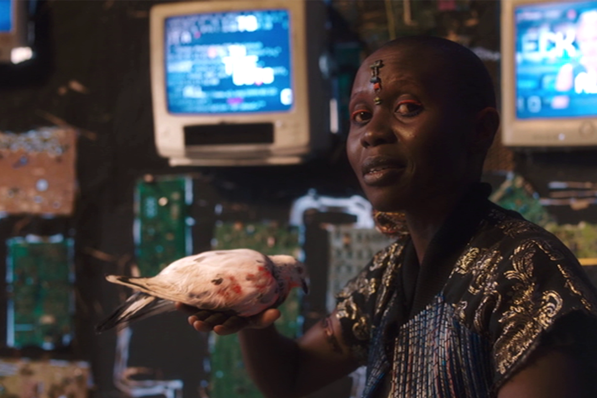 Still from Neptune Frost film, showing an African woman looking at camera, with a dove bird sitting on her hand and old fashioned computer monitors in the background