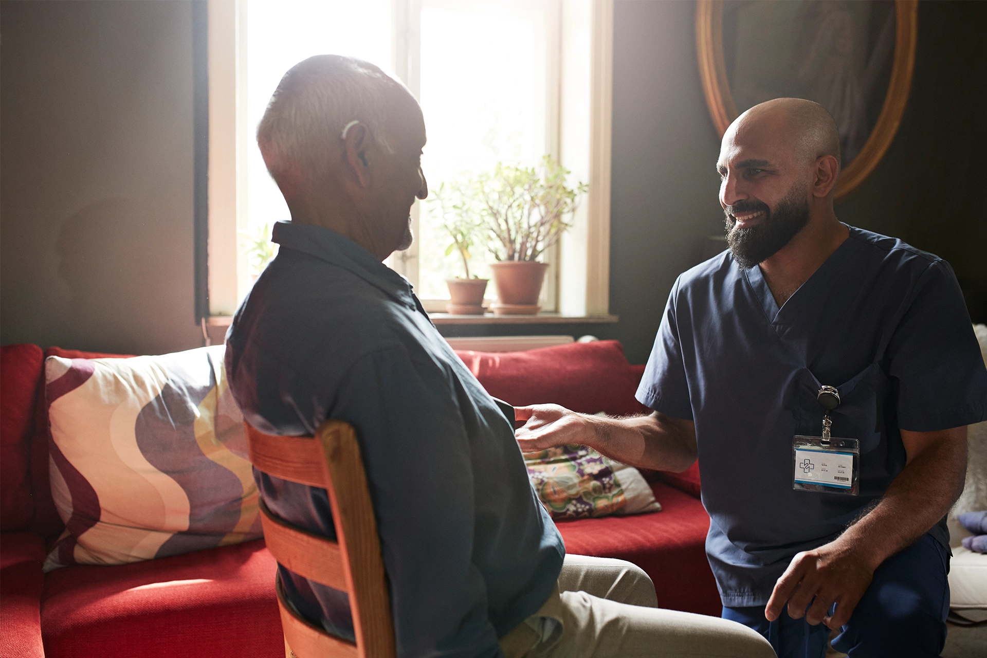 Smiling male nurse talking with senior man wearing hearing aid and sitting on chair in living room