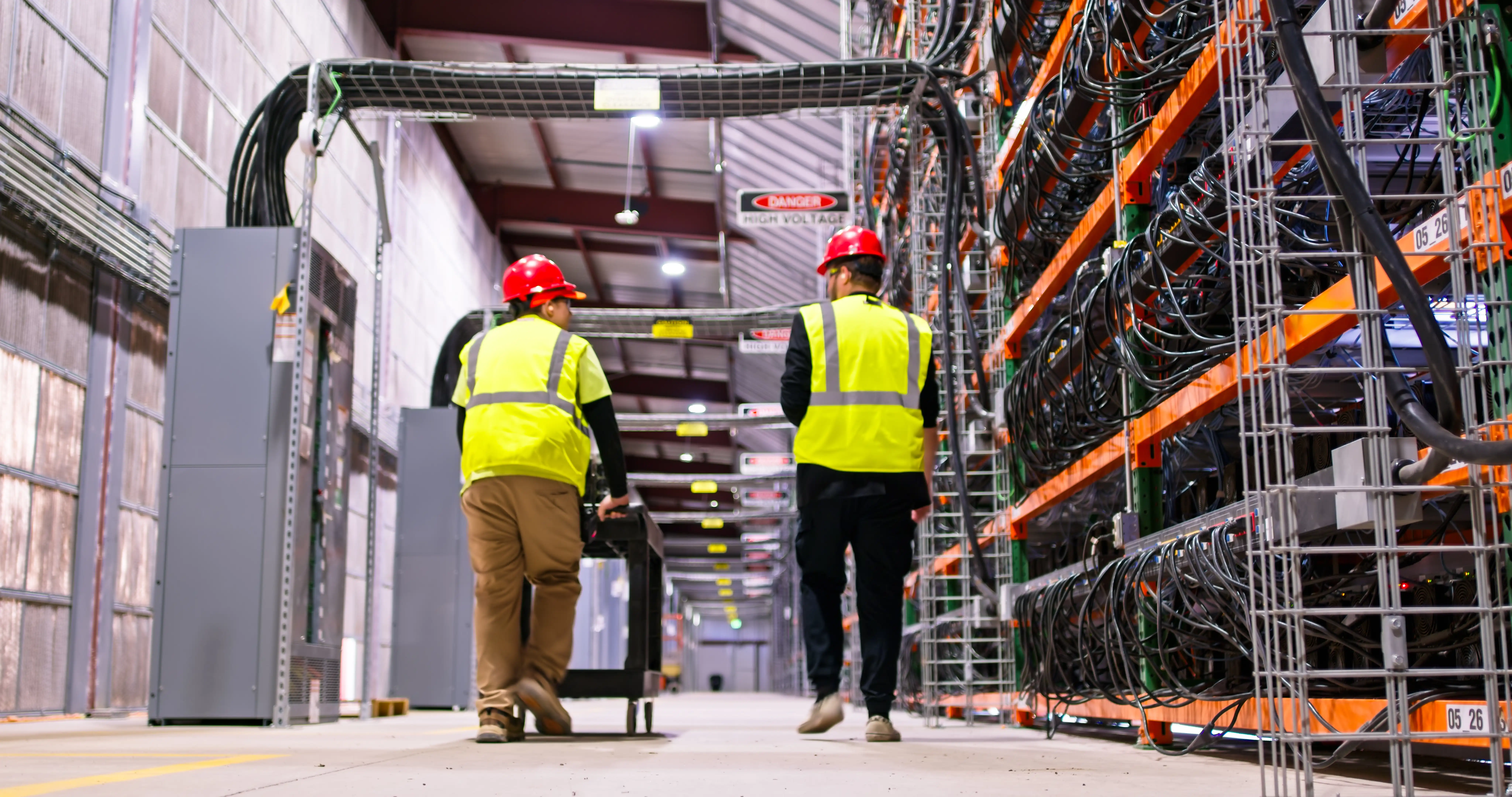 Two technicians wearing yellow high vis vests and red safety helmets walking through a large data centre, with tall shelves and cables spilling over the edge