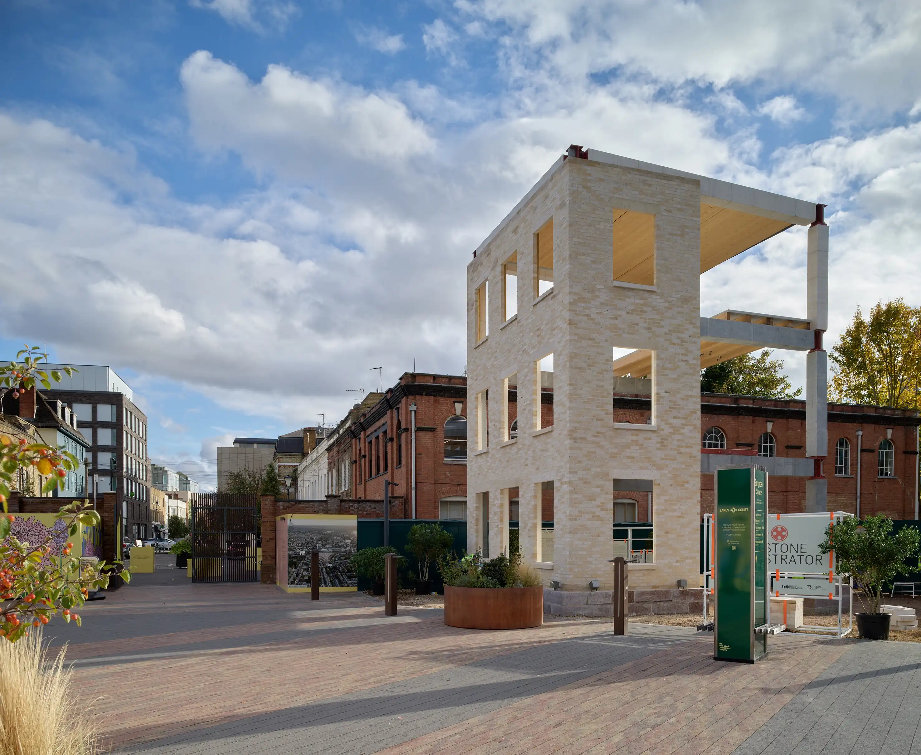 A three story frame for a building made primarily of stone, called the Stone Demonstrator, in an open square. There are signs around the building to explain its environmental impact and the sky is blue and full of clouds above it.