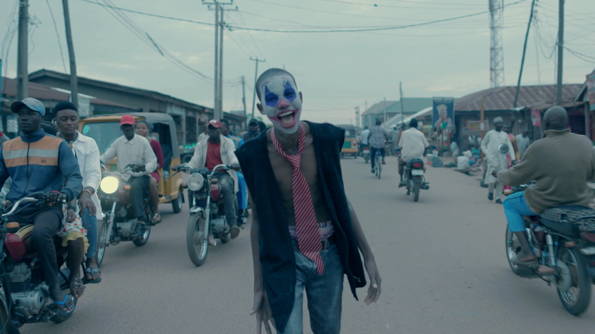 A film still of a young boy in clown make up and a tie walks the streets of Nigeria