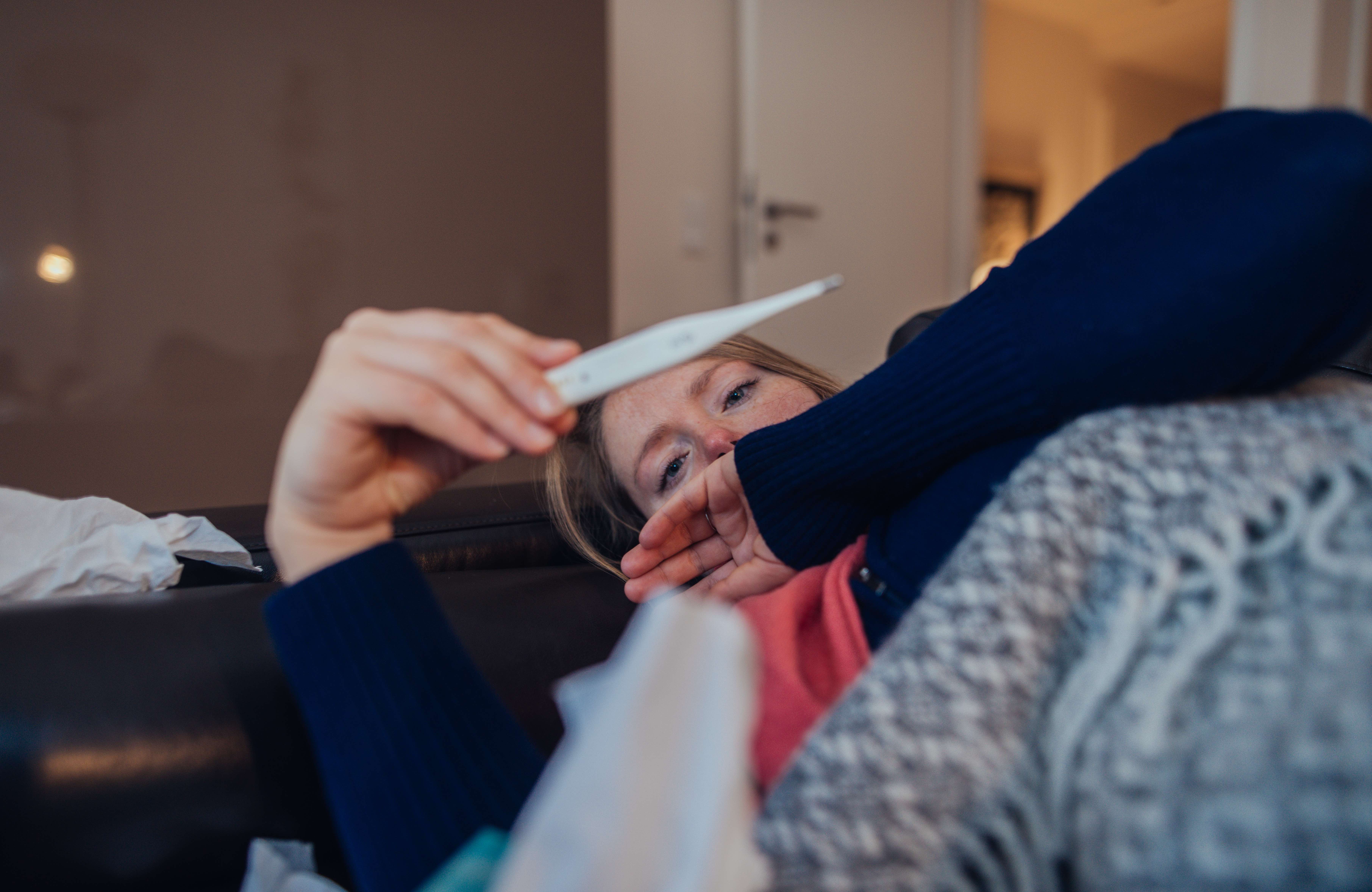 An ill woman on the couch looking at thermometre at home.