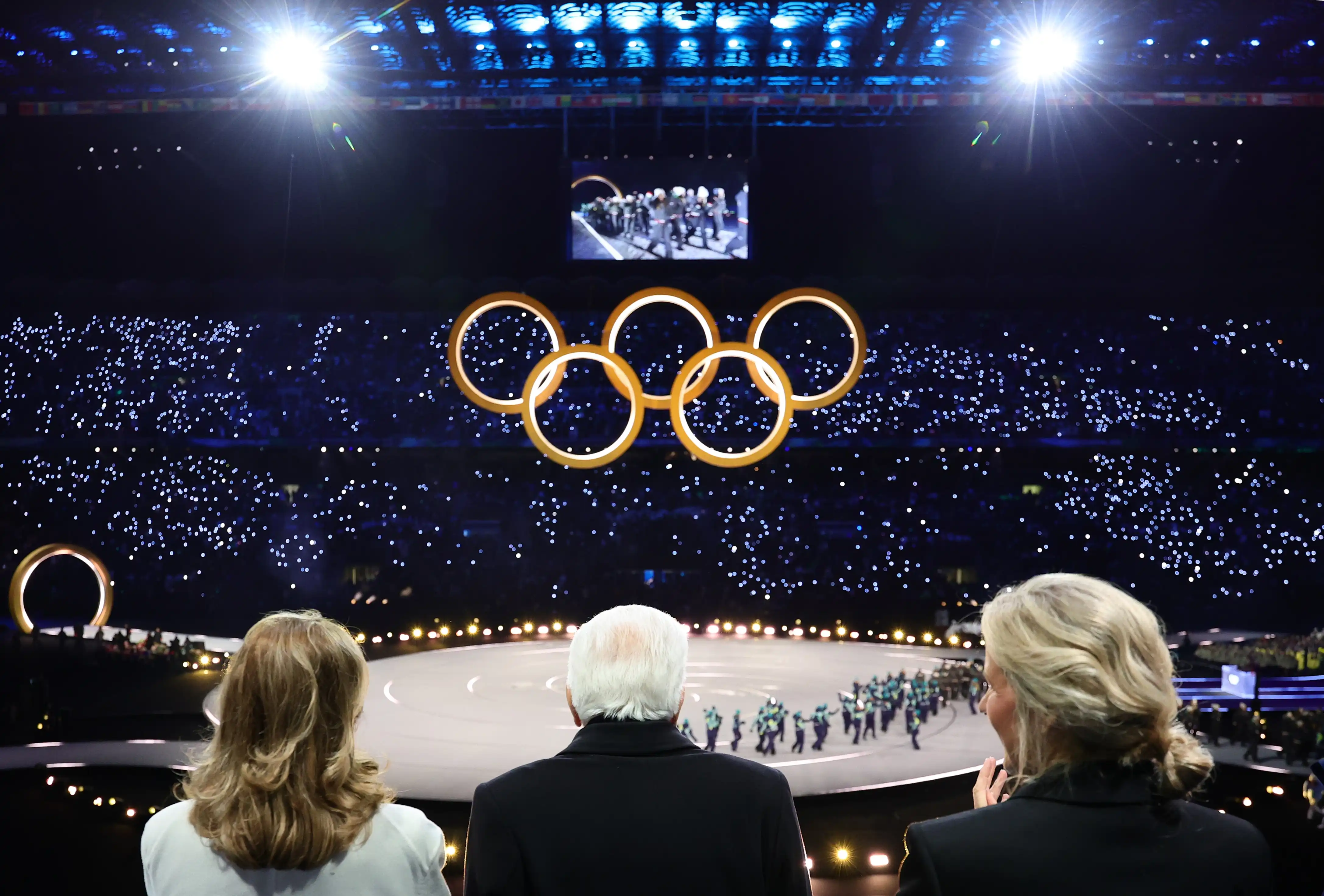 The backs of three heads look up at golden Olympic rings in a large sparkling stadium for the Opening Ceremony of the 2026 Winter Paralympic Games