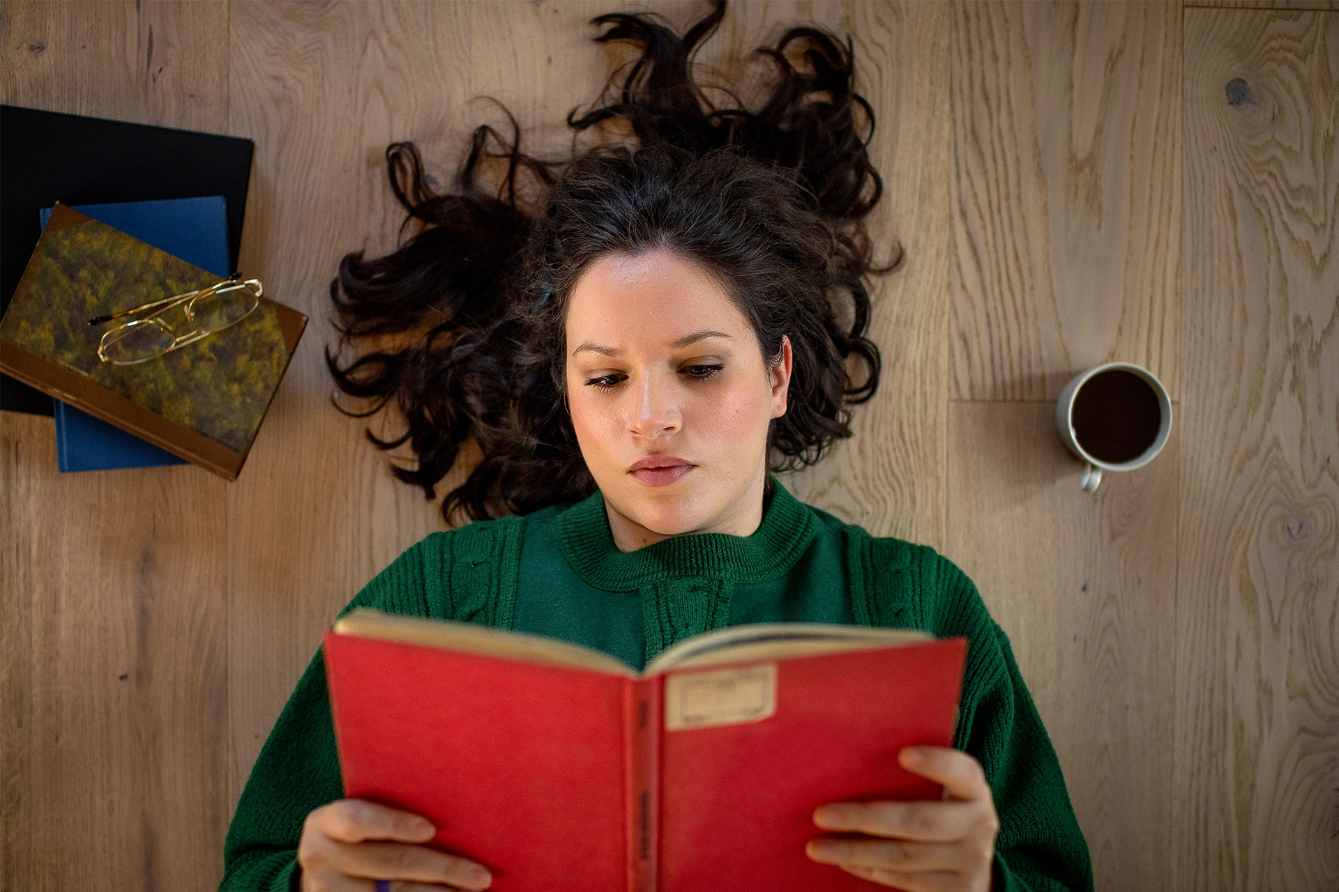 Woman with dark lying on the ground, reading a read hardback book with cup of coffee and reading glasses beside her