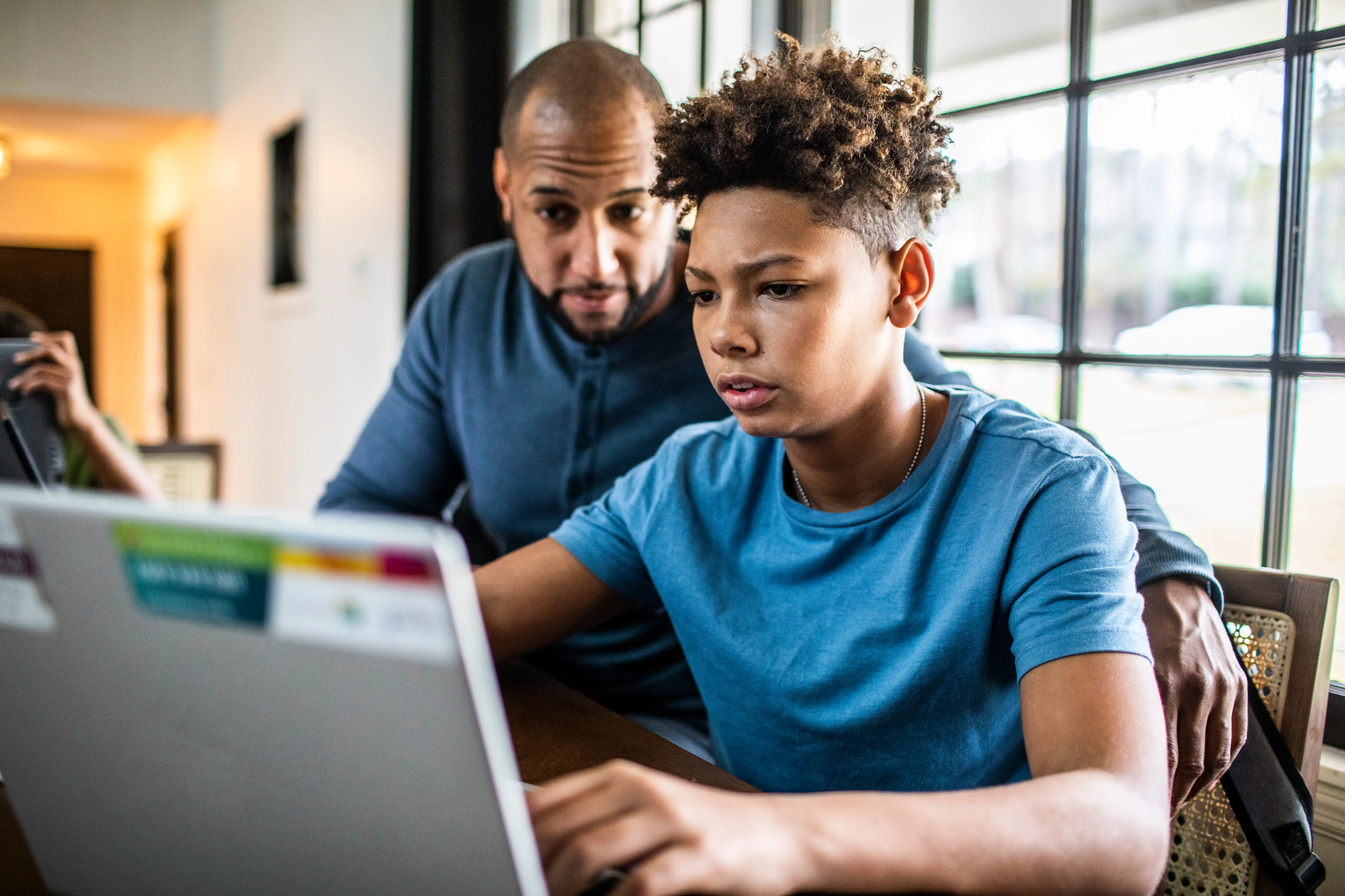 A father sits behind his son and helps him on a laptop