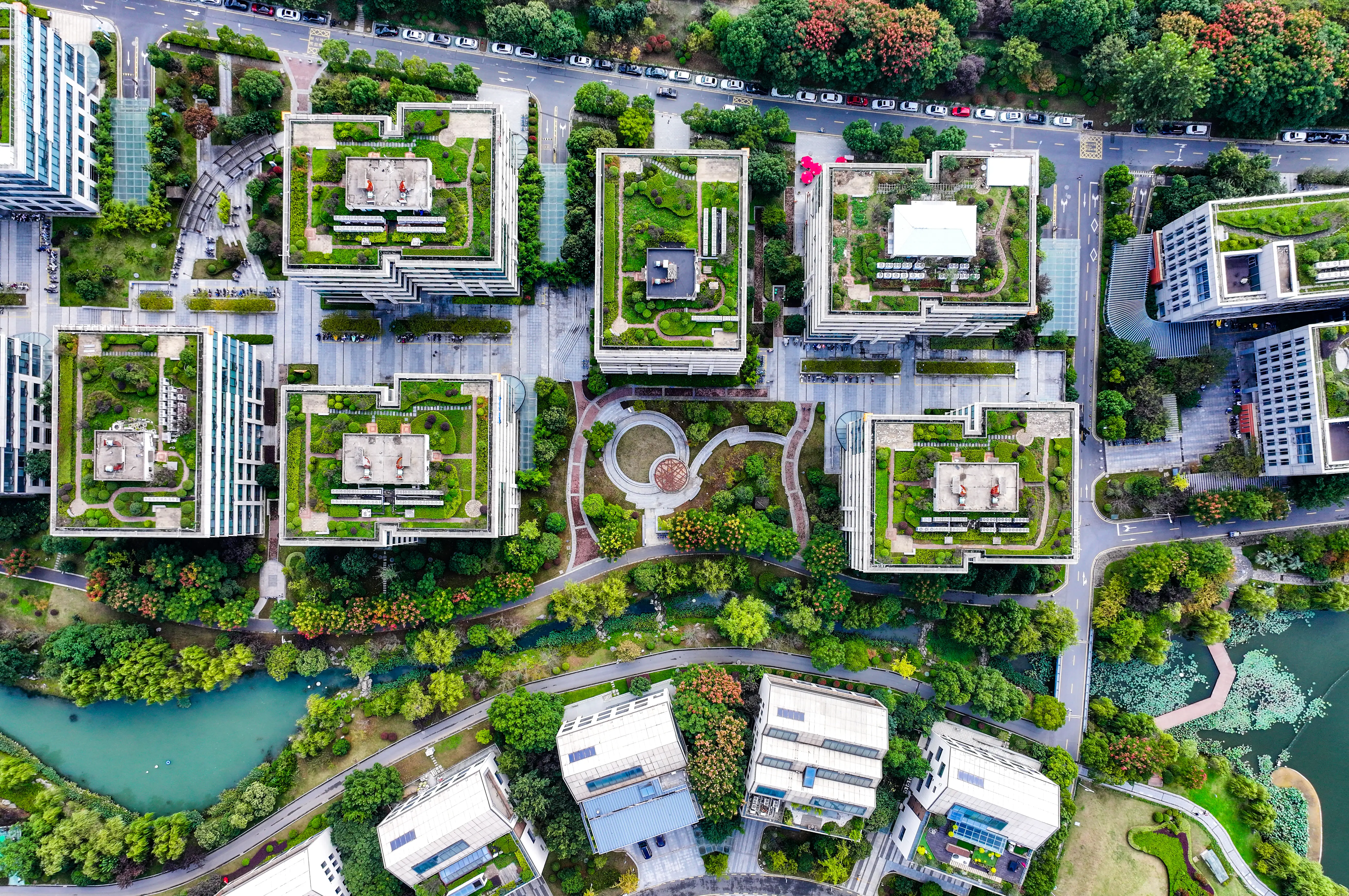 Aerial view looking directly down on a city district where office buildings with rooftop gardens surround a circular plaza, with tree-lined paths, a winding waterway and solar panels on nearby apartment blocks.