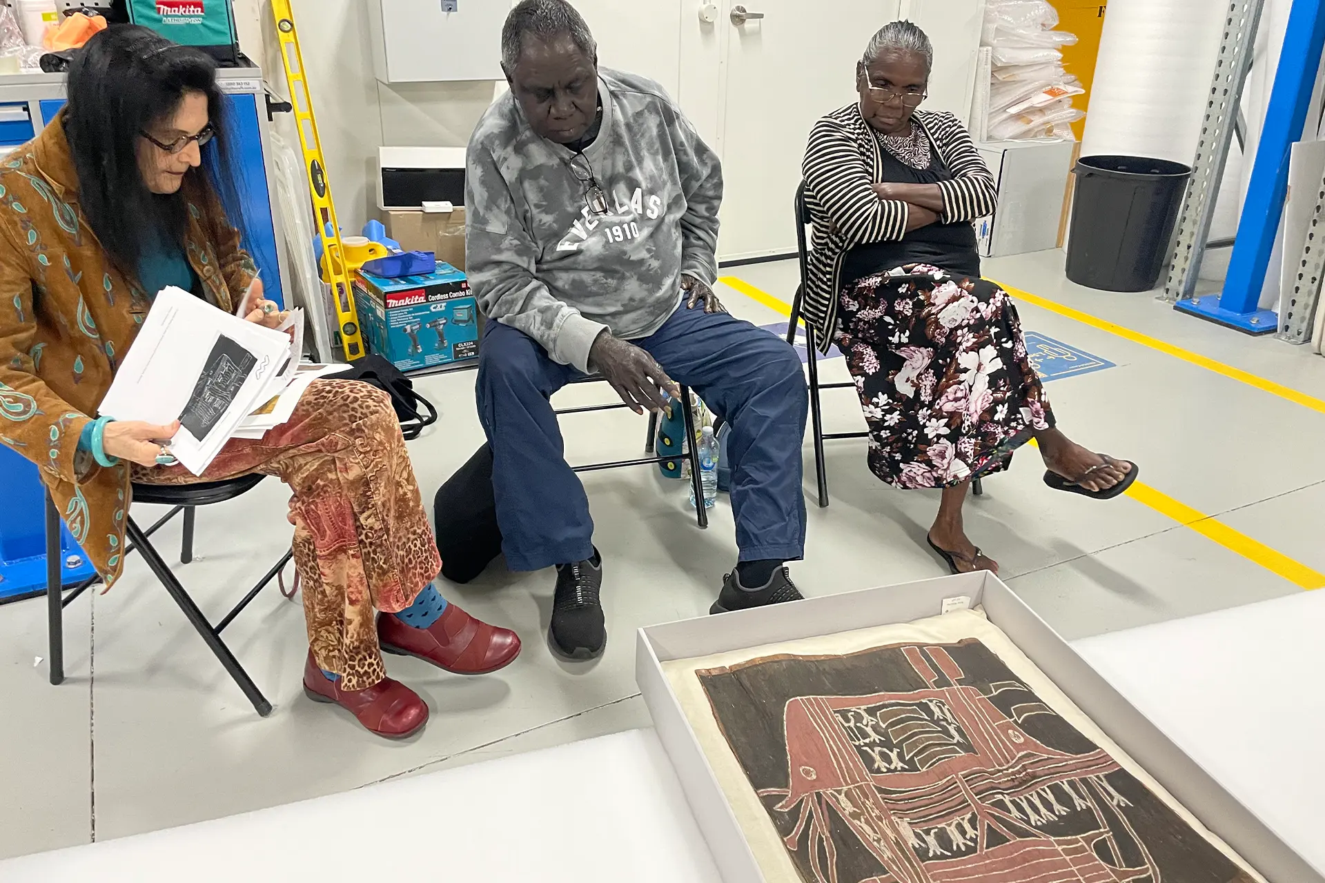 Three people sitting looking at an Aboriginal painting on the ground