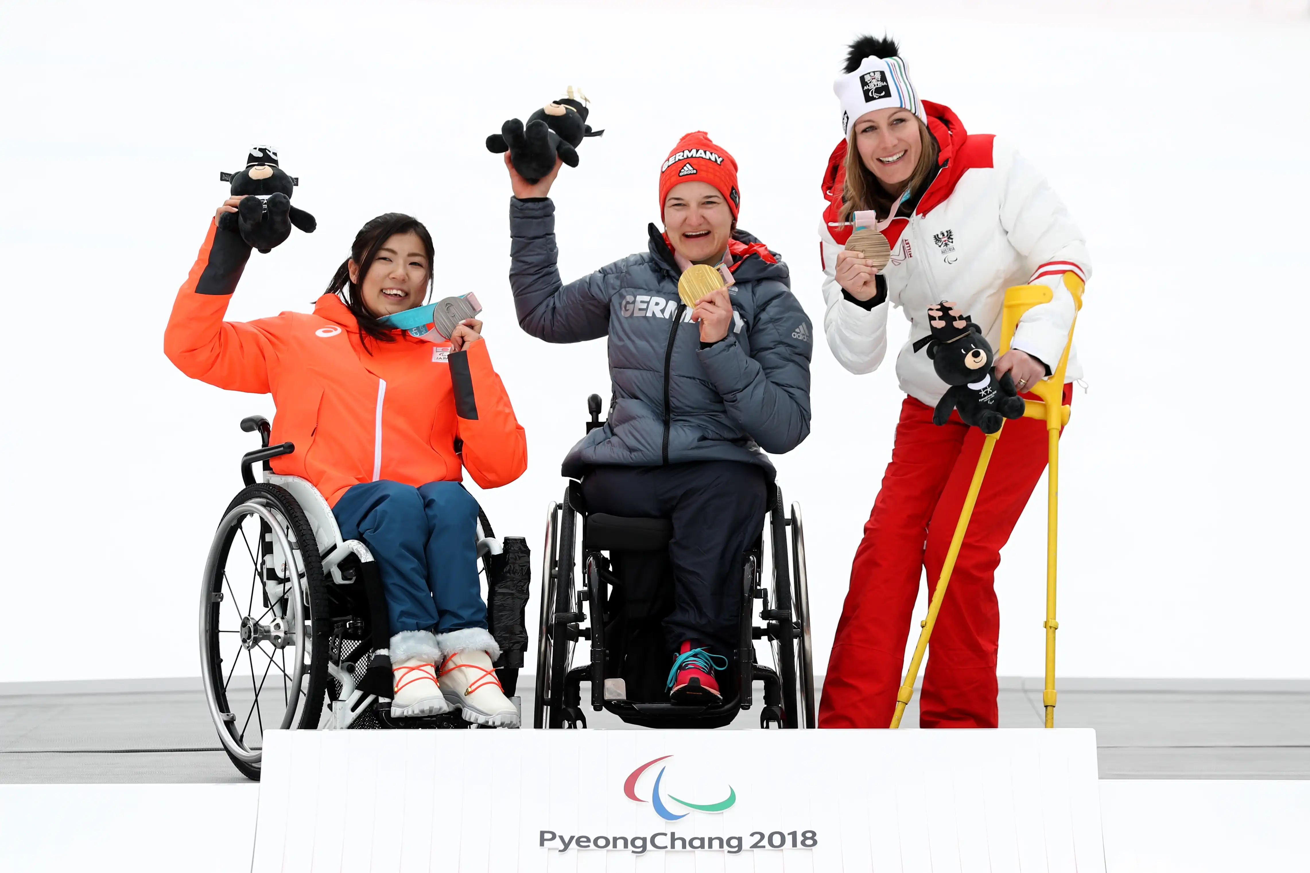 Three women in wheelchairs sit atop a podium in the snow showing their medals at the 2018 Winter Paralympics