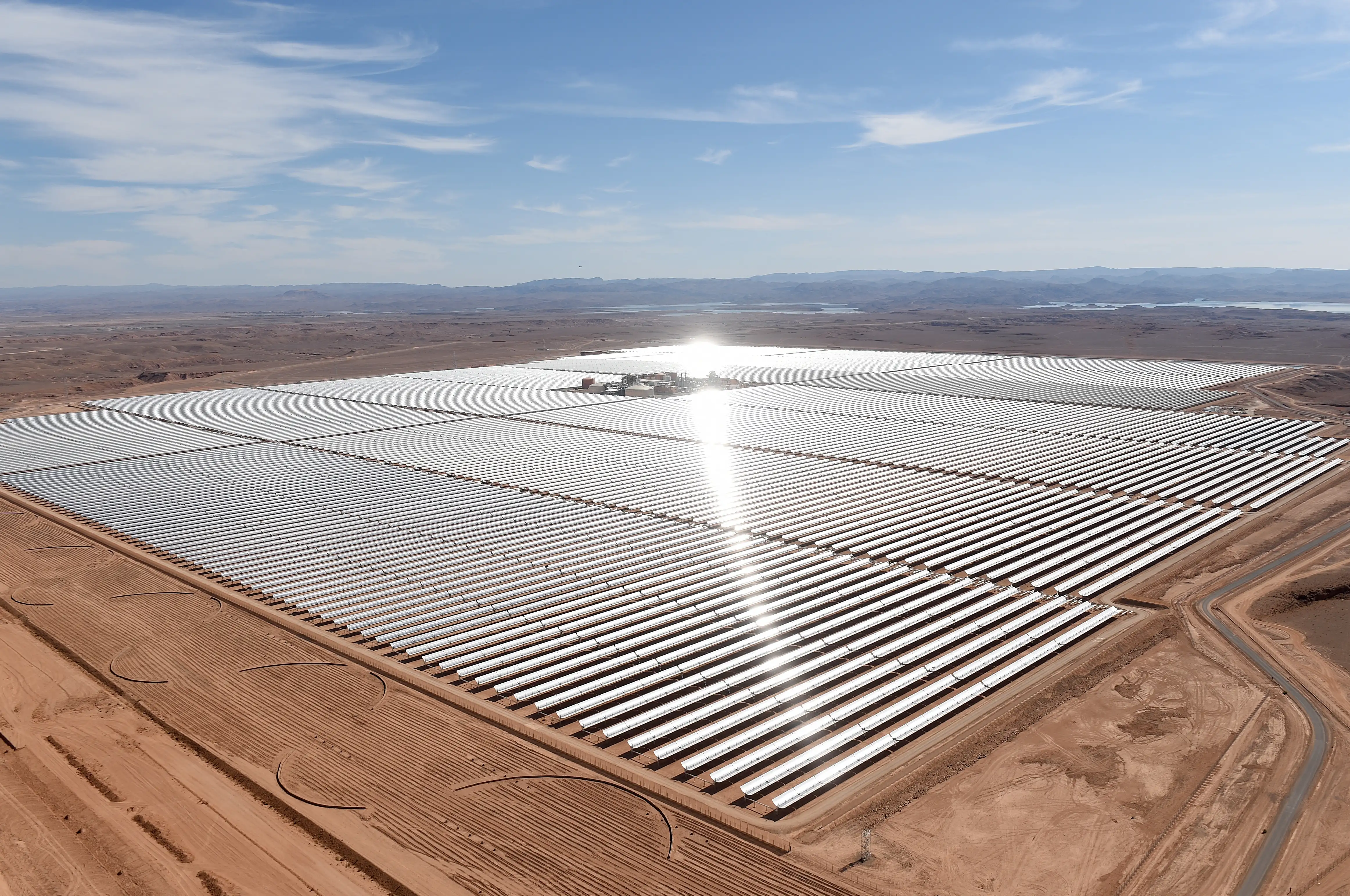 Aerial view of the vast Noor concentrated solar power plant in the Moroccan desert, with thousands of parallel rows of mirrors reflecting a bright blaze of sunlight and arid mountains on the horizon.