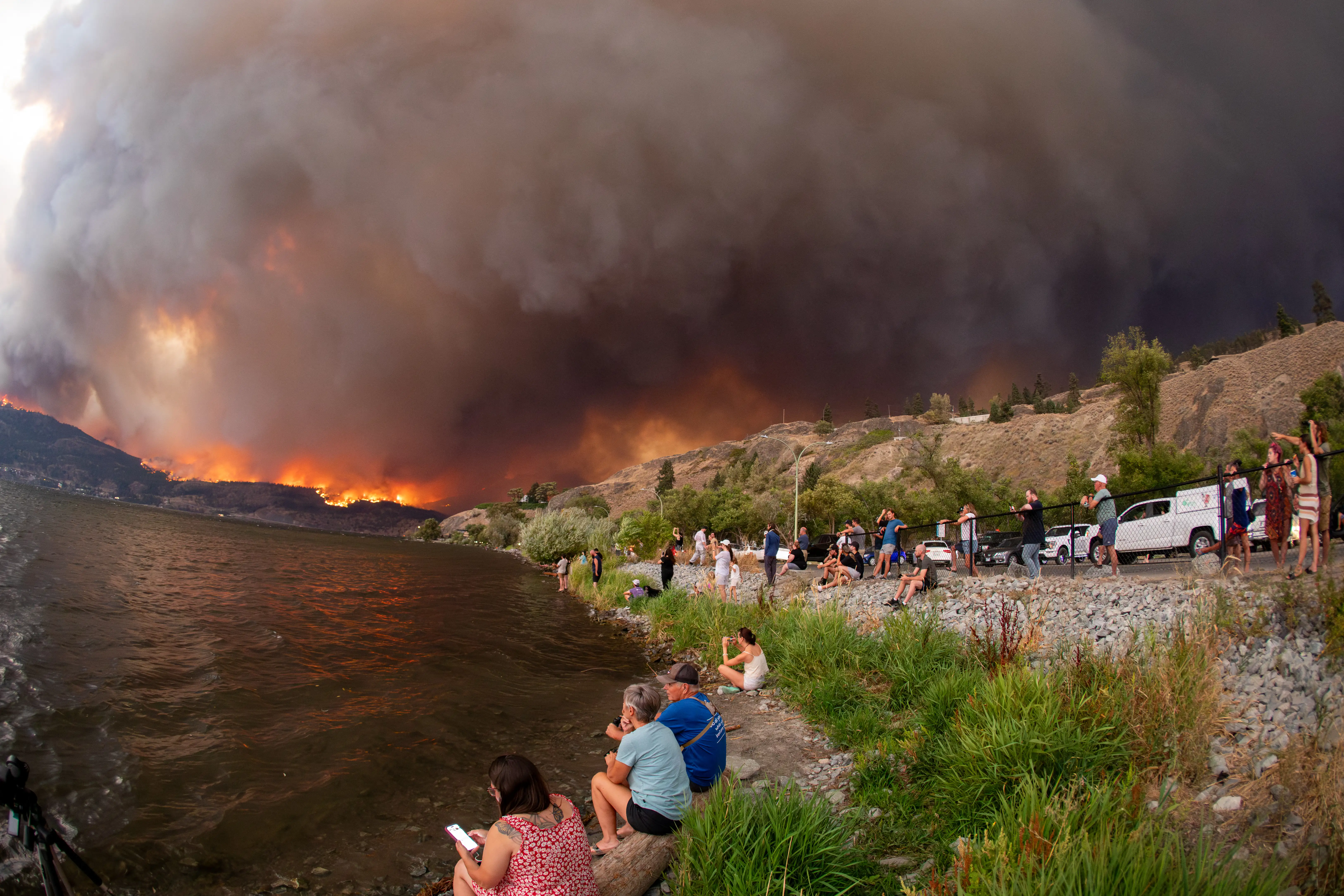 Smoke billows over a hill as people watch from a lake in Canada