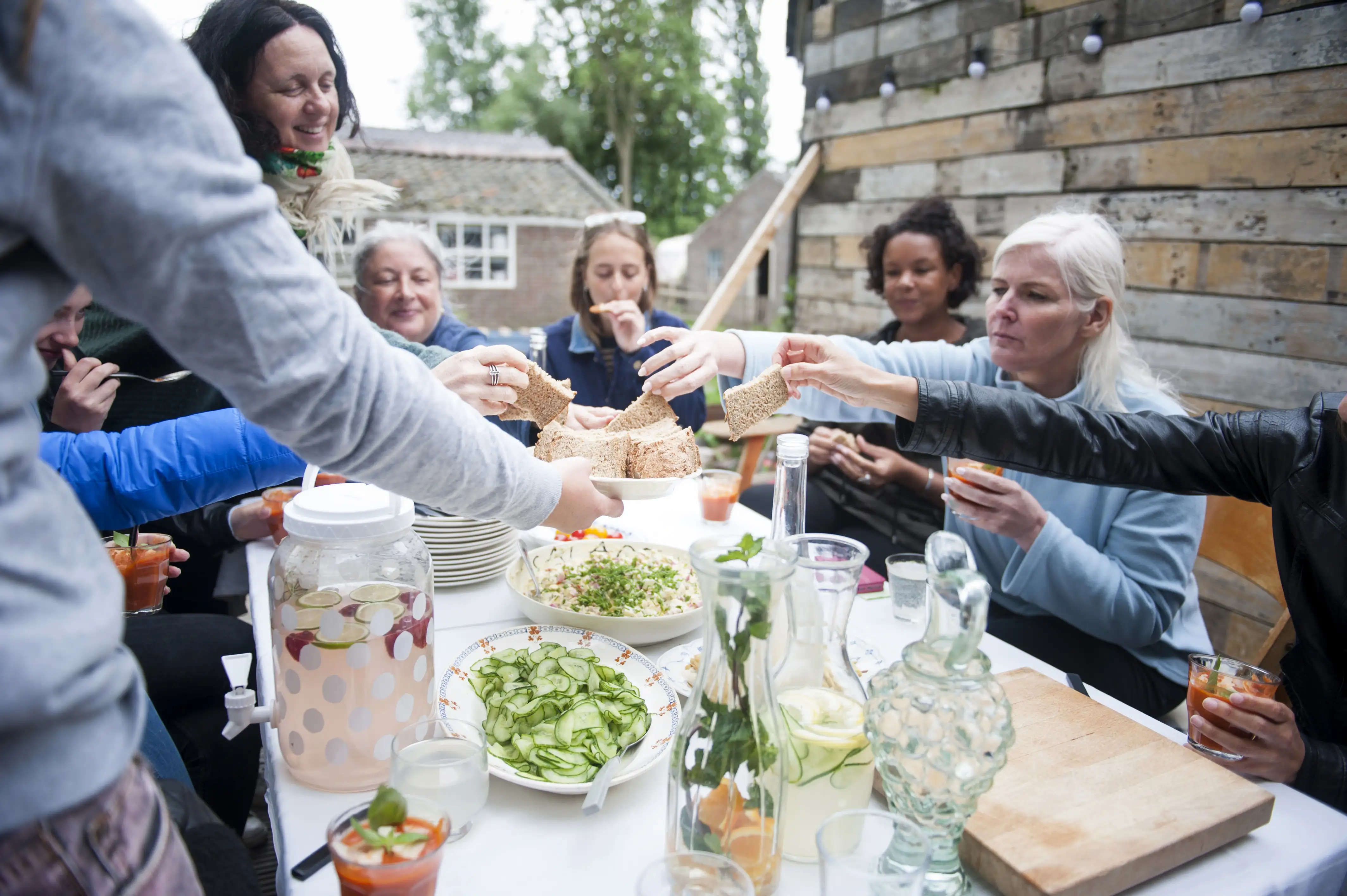 A group of women share a meal at a long outdoor table, passing pieces of bread across dishes of salad and jugs of homemade drinks, beside a wall of reclaimed timber.