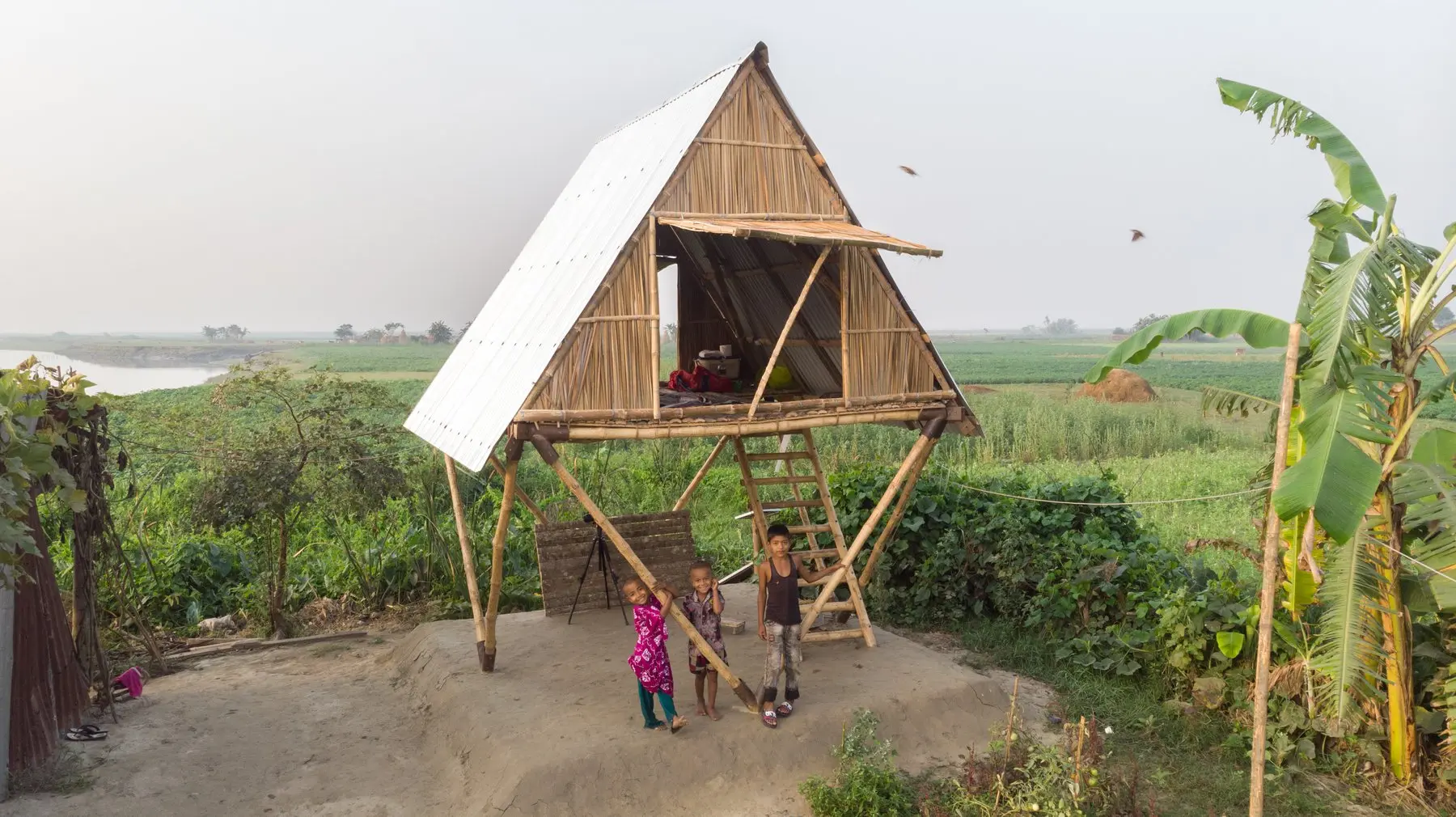Three young Bangladeshi children stand at the base of an elevated bamboo house, surrounded by green fields and a river