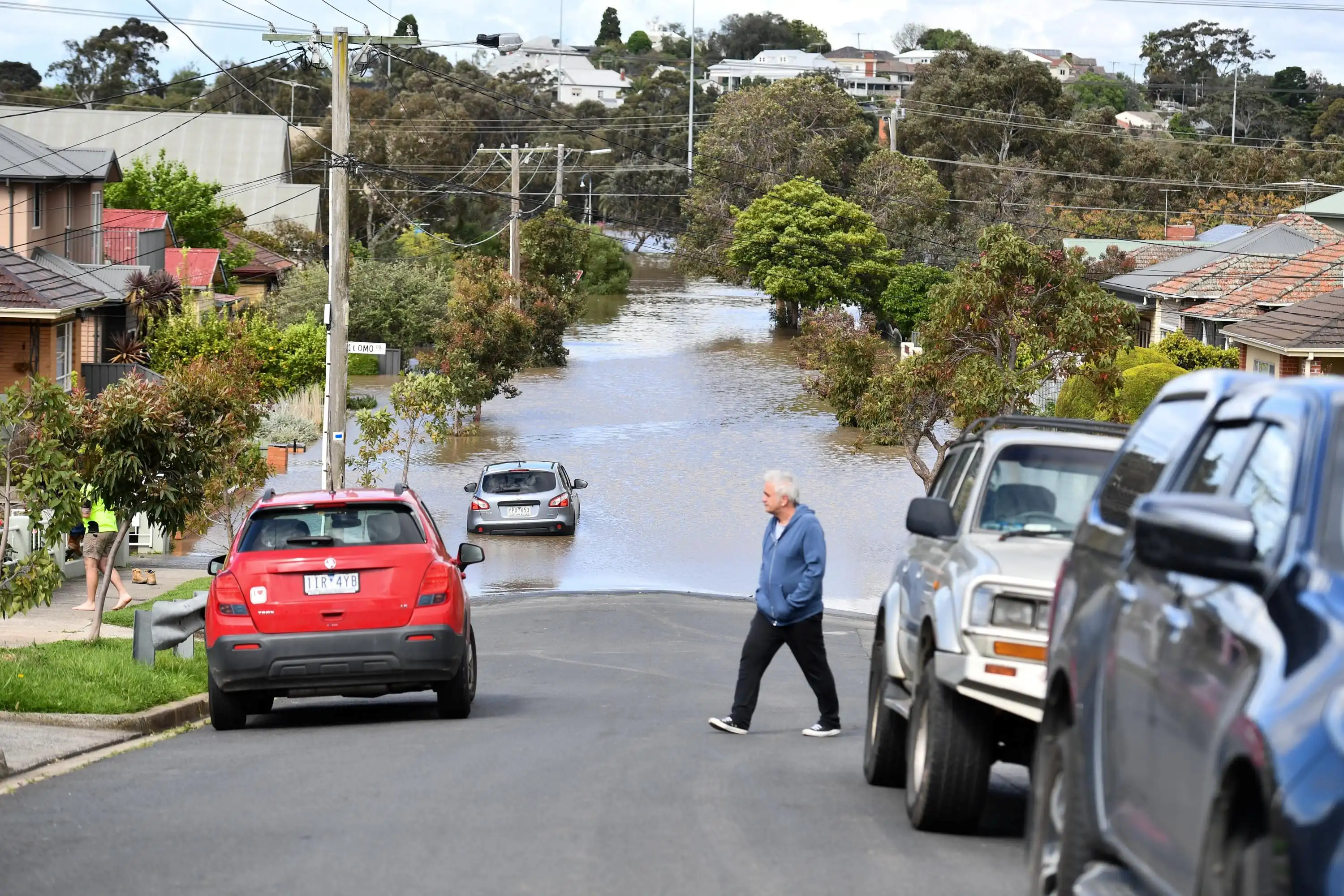 A man walks across a steep road as the lower part of it is flooded up to houses