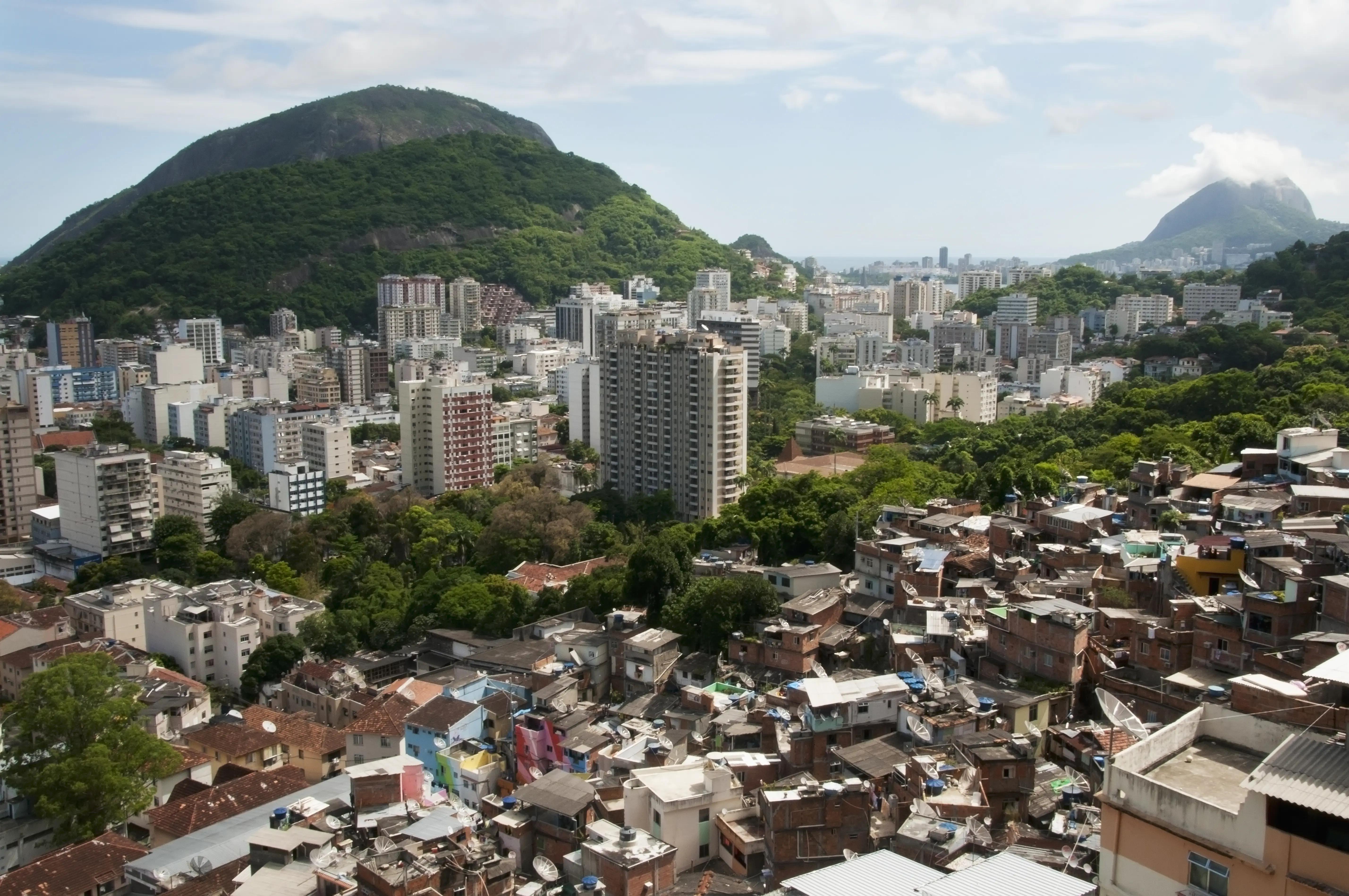 View across Rio de Janeiro showing a densely packed hillside favela of brick and tin-roofed homes in the foreground, separated by a strip of trees from white high-rise apartment towers, with forested mountains behind.