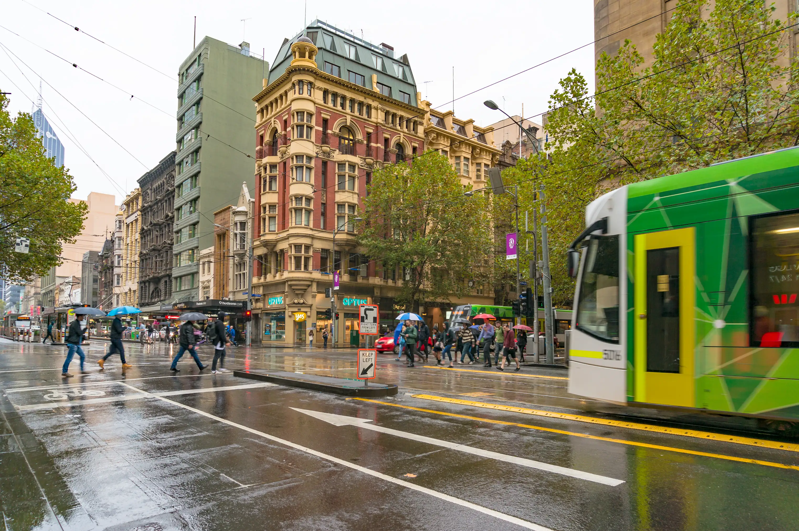 Pedestrians with umbrellas walk across a rainy road in front of a Melbourne tram