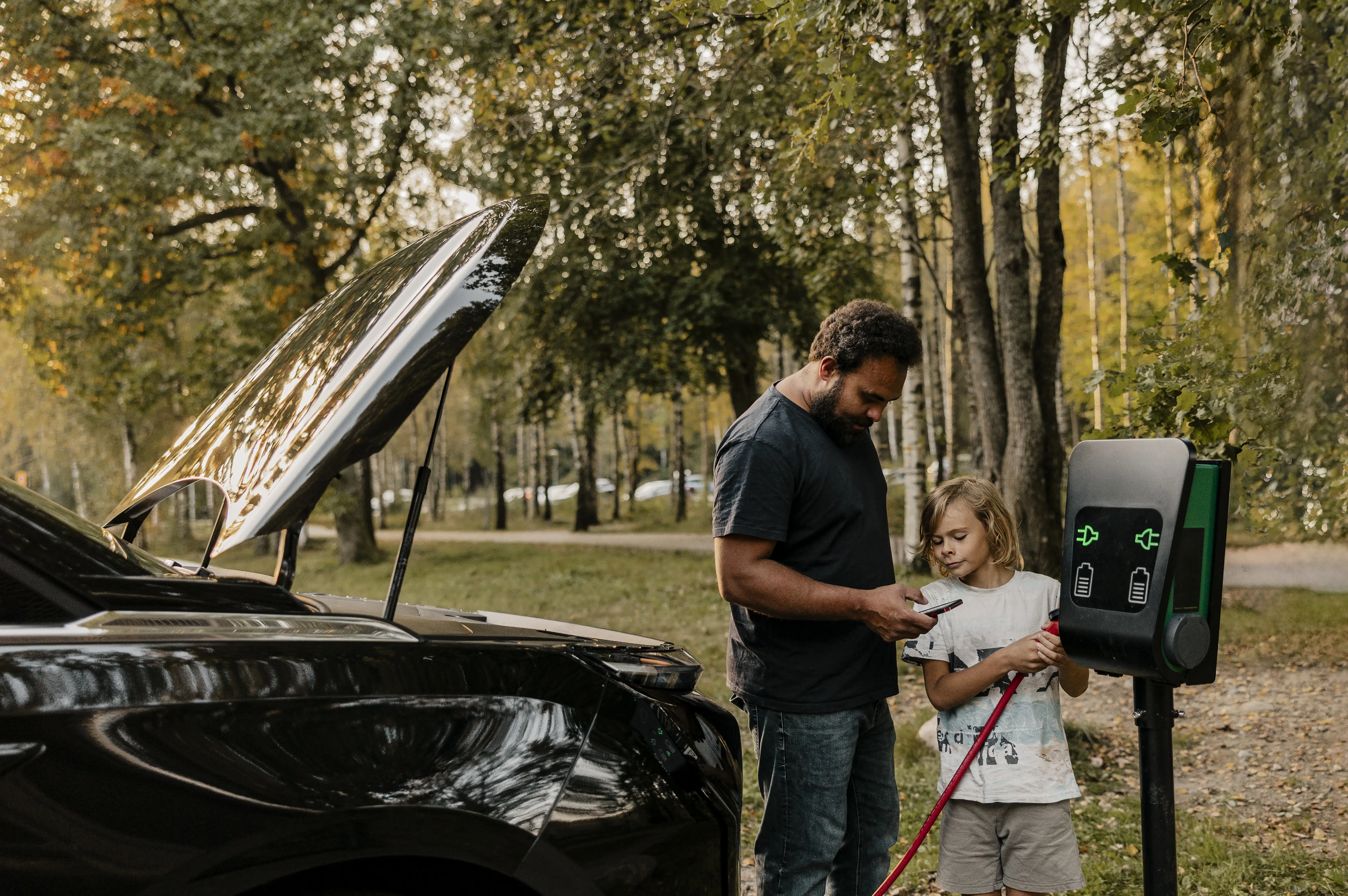 Father using smart phone while son charging electric car near forest